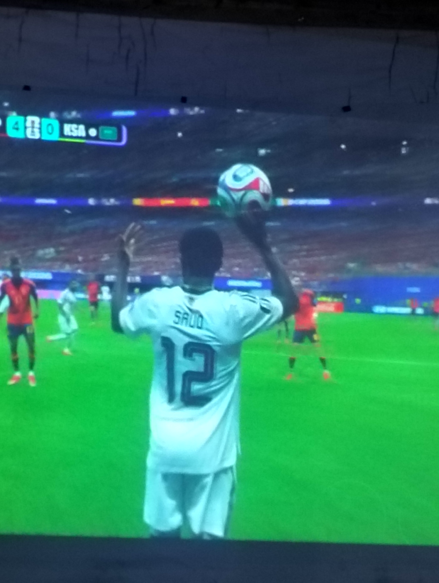 A soccer player in a white jersey with the number 12 on his back and the name "SAUD" above it is holding a soccer ball above his head on a green soccer field. He is facing away from the camera, and the ball is in his right hand. The player is wearing white shorts. In the background, there are other soccer players in red jerseys. There is a scoreboard visible in the upper left corner of the image, showing "4-0" and "KSA". The image appears to be taken from a screen, possibly a television, as there are some reflections and a somewhat grainy quality to the image. The location in the city of Payagaboh, Indonesia is not visible or discernible from the provided image.
