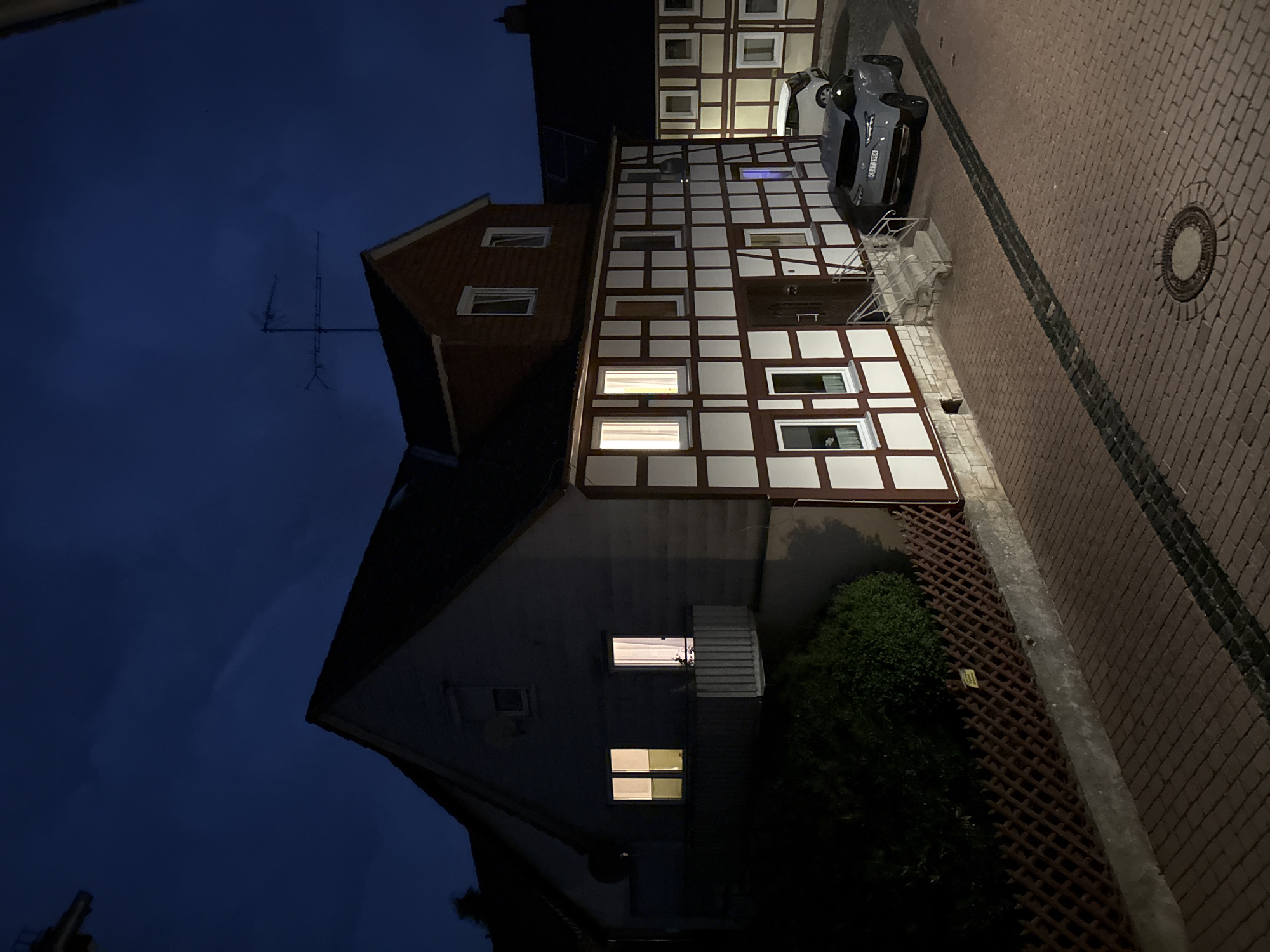 In an outdoor scene captured at dusk, a multi-story building with a dark tiled roof dominates the frame. The building exhibits traditional German Fachwerk architecture, characterized by exposed timber framing painted in a dark reddish-brown color against white infill walls. Several windows are visible, some illuminated from within, casting a warm glow. A balcony with a white railing extends from the left side of the building. Adjacent to the main structure, another building with similar architectural features is partially visible on the right, also featuring illuminated windows.
In the foreground, a paved driveway or street is visible, constructed from reddish-brown brick pavers. A dark gray modern car is parked on the right side of the driveway, its taillights illuminated. Behind it, a white SUV is also parked. A low, dark brown lattice fence runs along the bottom left of the image, separating the paved area from a dark, overgrown patch of greenery. A single yellow sign is affixed to the fence. The sky above is a deep, dark blue, suggesting twilight or night. An antenna is mounted on the rooftop of the main building.
