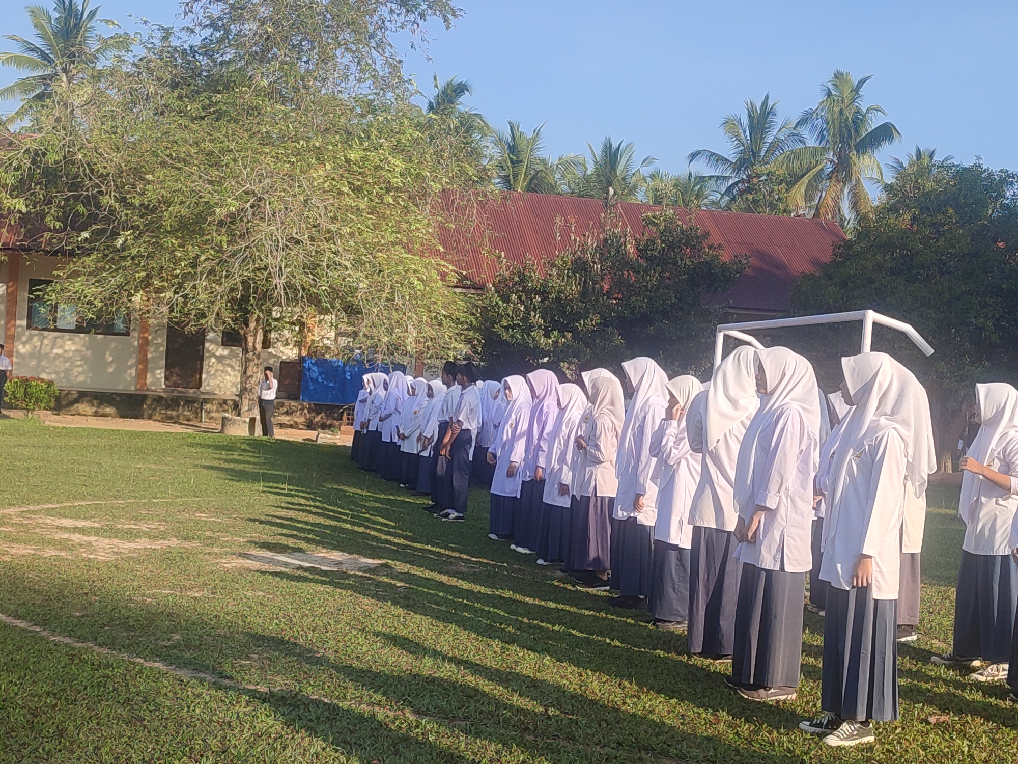 A group of people, primarily women wearing hijabs and long skirts, are standing in a line on a grassy field. They are dressed in white shirts and dark skirts. The field is situated in front of a building with a red roof. The location appears to be Blangriek, Indonesia. A soccer goalpost is visible. There is at least one male individual in the background. The general environment consists of greenery and trees.