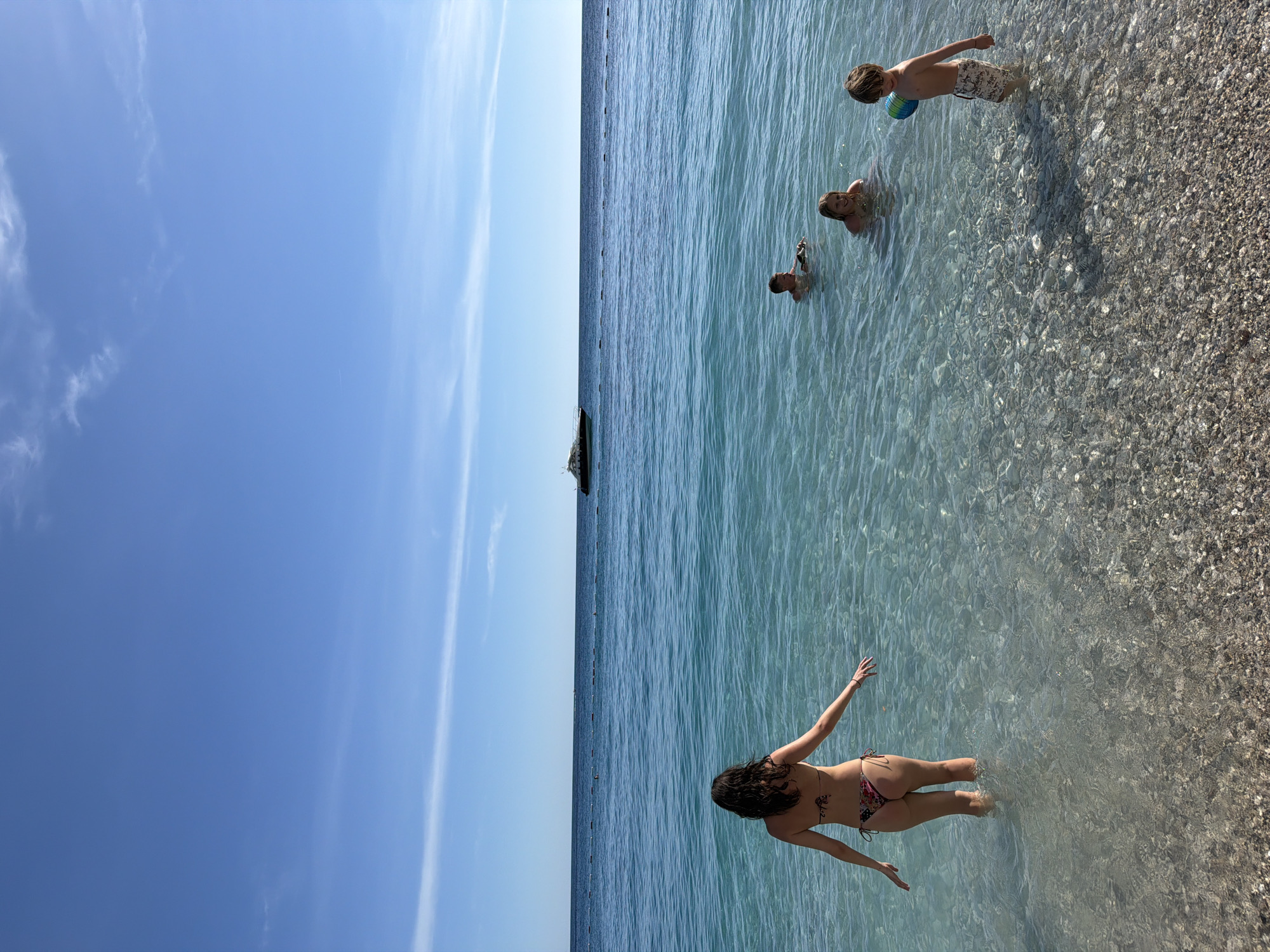 The image shows a beach scene in Sveti Stefan, Montenegro. The sky is clear and blue. The water is clear, with a rocky shoreline. In the foreground, a woman in a bikini is standing in the water with her arms outstretched. Further out, three people, including a boy with a ball, are playing in the water. A structure is visible on the horizon. The beach appears pebbly.