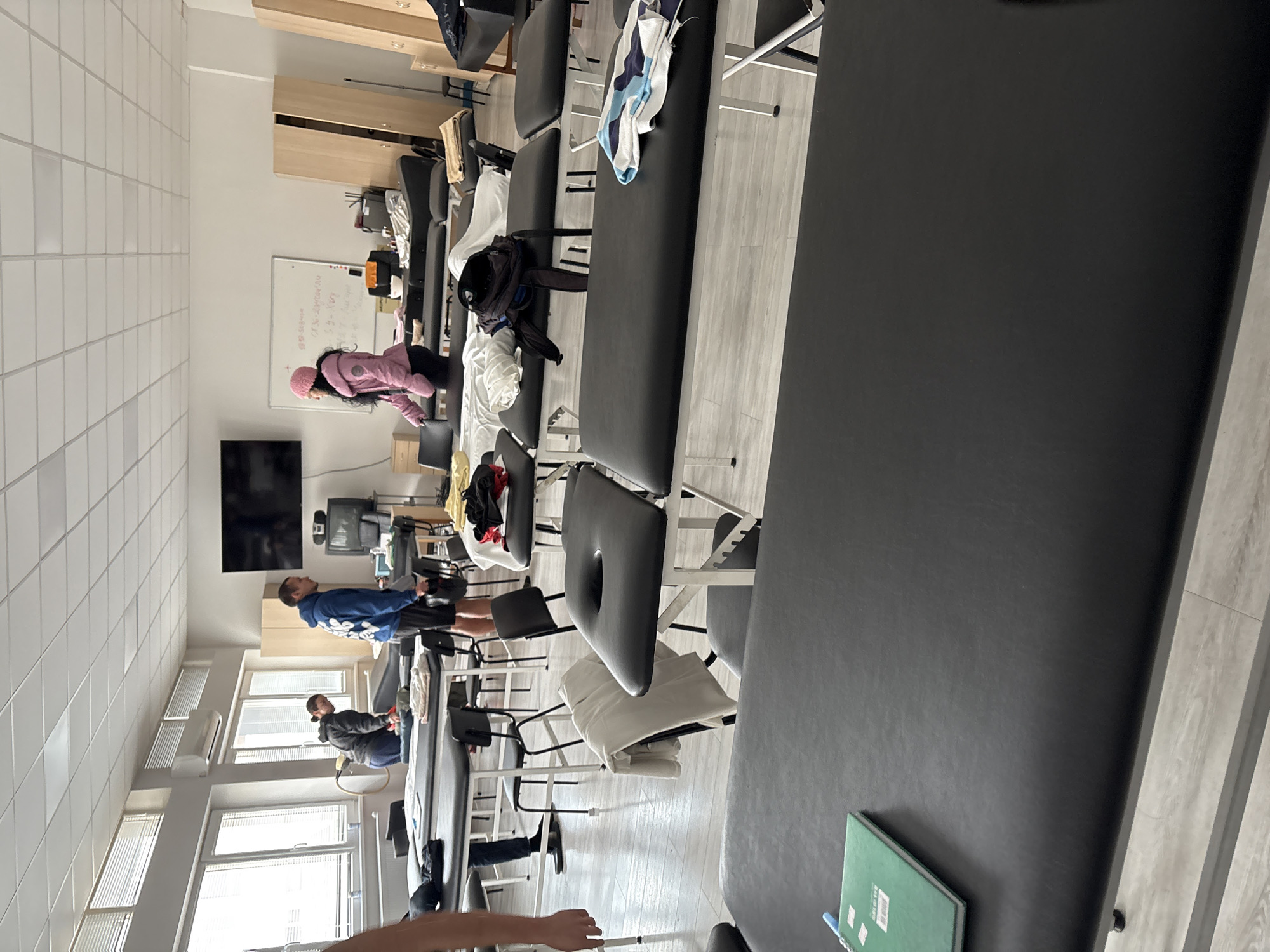 The photograph shows a training room, possibly for medical or physiotherapy students, in Sofia, Bulgaria. The room is equipped with multiple treatment tables. Several people are present. A person in a blue shirt is standing near the window, possibly performing a stretching exercise. Another person in a grey jacket is facing the same direction. Further down the line of tables, a person in a pink coat and hat is lying on one of the tables. The room contains a whiteboard with writing on it, storage cabinets, a television screen on the wall, and various medical devices. The flooring appears to be a light-colored wood or laminate. Natural light is entering through the windows. The treatment tables are covered in black vinyl.
