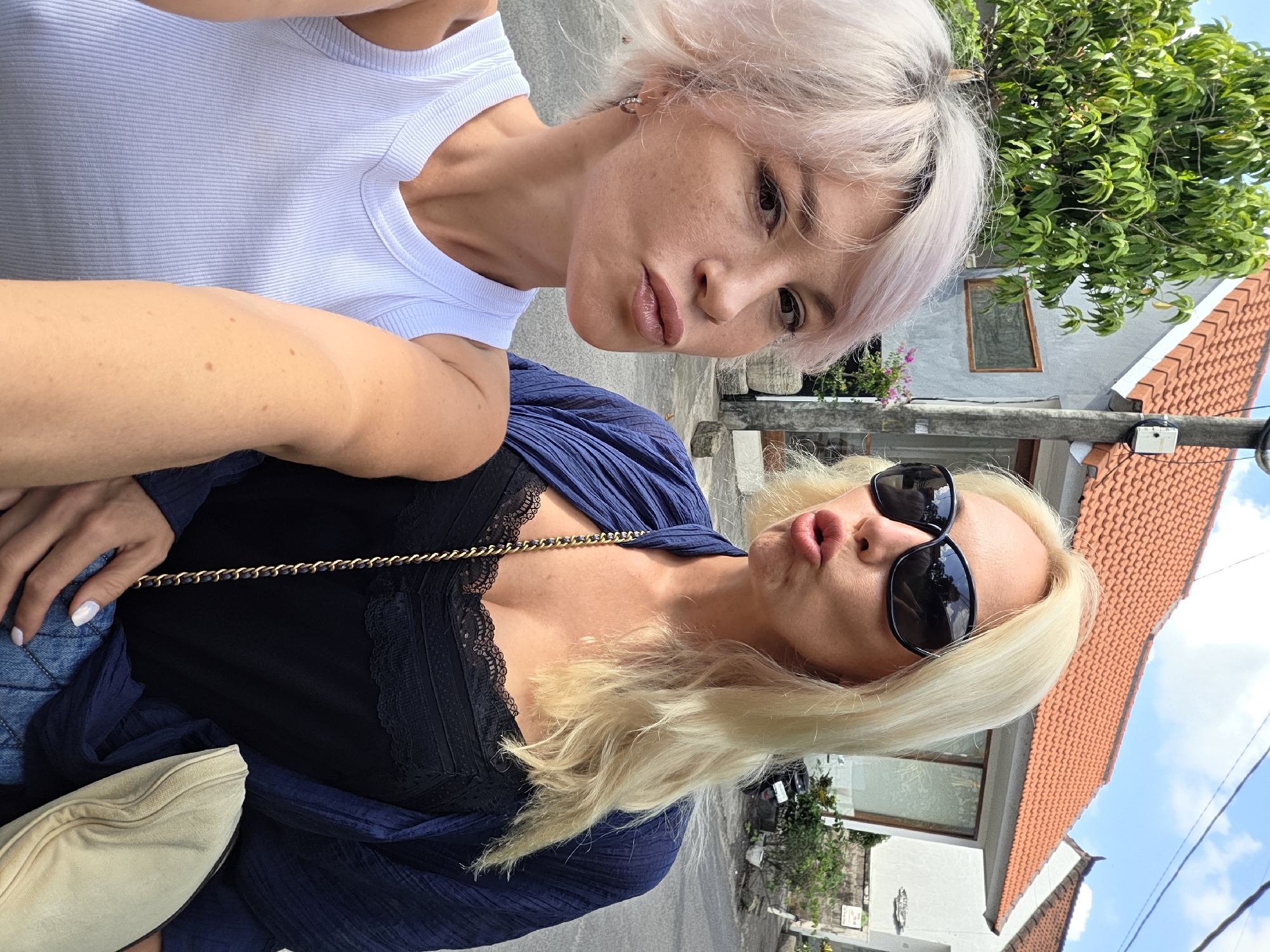 Two women are taking a selfie outdoors in front of a building with an orange tiled roof. The woman on the left has short, light pink hair and is wearing a white tank top. She is making a pouting face. The woman on the right has long blonde hair and is wearing large black sunglasses, a black lace-trimmed tank top, and a dark blue open cardigan. She is also making a pouting face. They are both in a street, with a tree and a power pole visible in the background, suggesting an urban or suburban environment in North Kuta, Indonesia. The overall scene is casual and appears to be during the daytime under a bright sky.