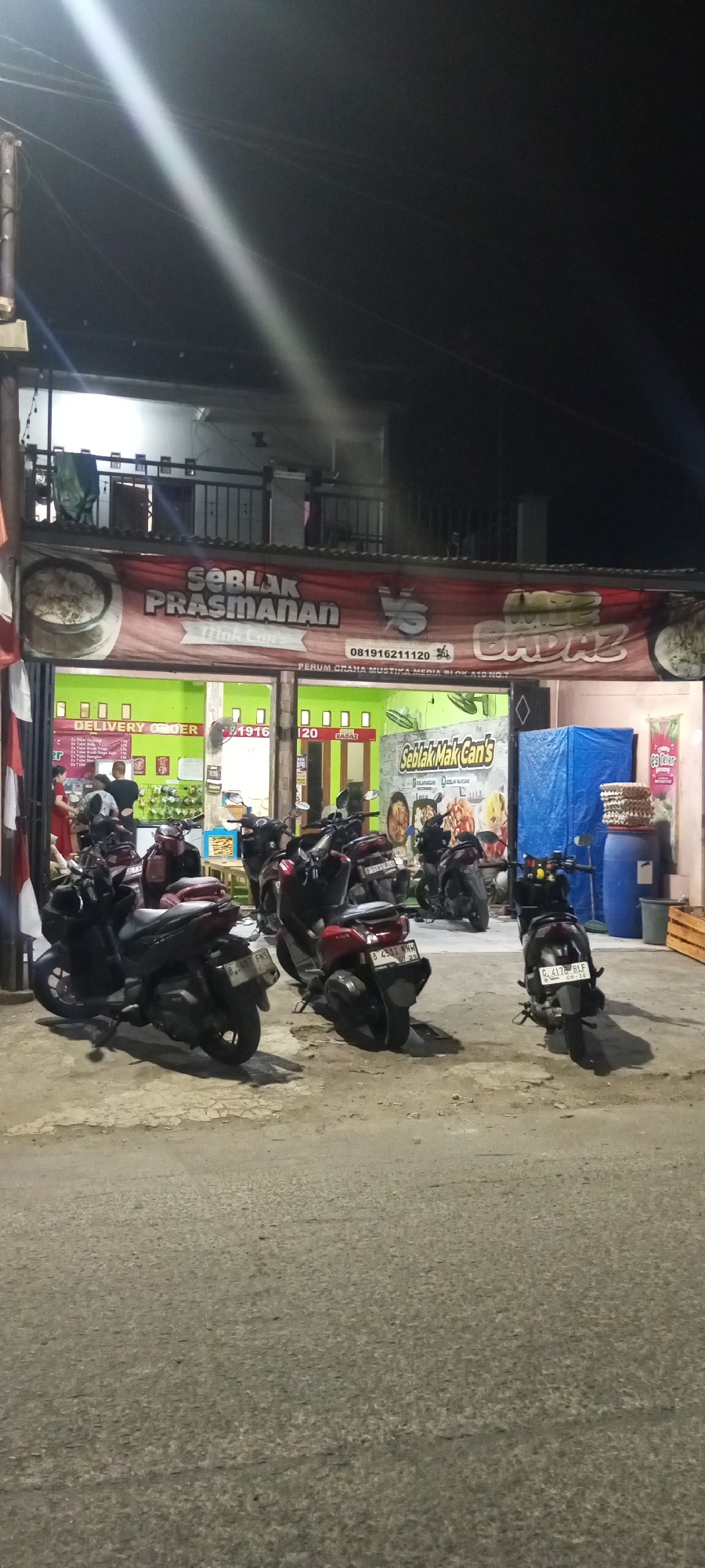 The image depicts the exterior of a food establishment named "Seblak Prasmanan" located in Setu, Indonesia. The establishment appears to be a small shop with a red banner displaying its name and a phone number, "081916211120". Below the banner, the shop's interior is visible, featuring green walls and what appears to be a menu or promotional material. A sign with "DELIVERY ORDER" is also present. Several motorcycles are parked in front of the shop, suggesting it is a popular spot or a location for delivery services. The lighting indicates it is nighttime, with a bright light beam cutting across the upper portion of the image, possibly from a street light or a strong spotlight. The overall environment suggests a typical street scene in a residential or commercial area of Setu at night.