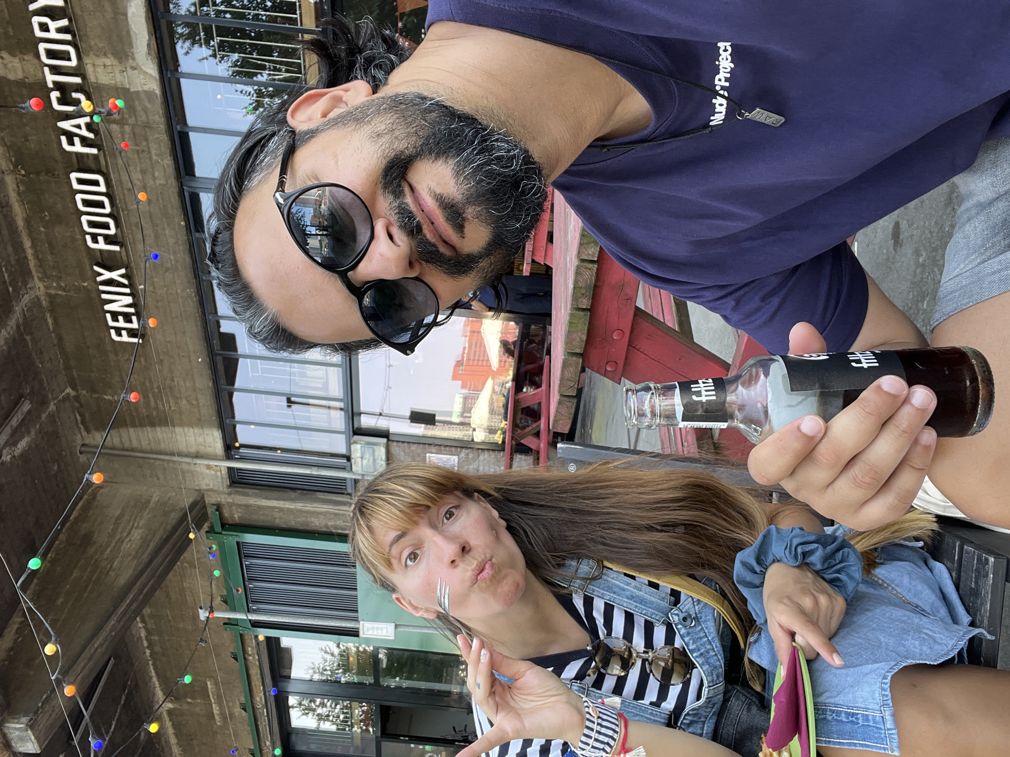 This media file shows two people sitting at an outdoor table, likely at a restaurant or cafe, with a building and some trees in the background. The setting appears to be Rotterdam, Netherlands. The time of day seems to be daytime, given the lighting.
The person in the foreground, on the left, is a man with dark hair and a beard, wearing sunglasses and a dark blue t-shirt with "Nudx Project" and a small silver pendant visible on a necklace. He is holding a dark glass bottle with a label that reads "fritz".
The person in the foreground, on the right, is a woman with long blonde hair, wearing a denim jacket over a striped t-shirt. She is holding a fork and has a playful expression on her face, pursing her lips. She is also wearing sunglasses clipped to her shirt and a blue scrunchie on her wrist.
On the building behind the man, there is white text that reads "FENIX FOOD FACTORY". String lights with colored bulbs are visible, adding a festive touch to the scene. The overall mood appears relaxed and casual.