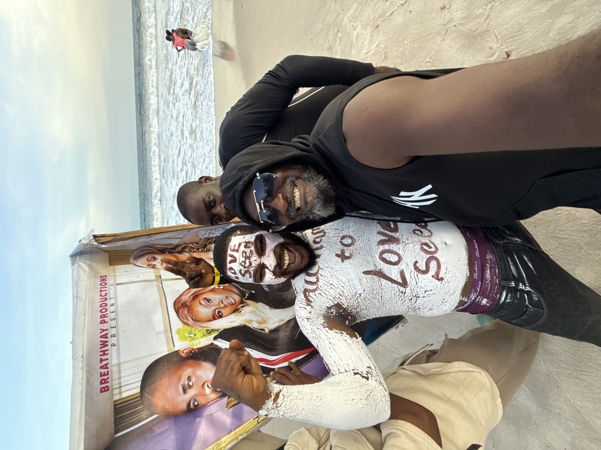 The image shows three Black men lying on a beach in Mopo Onijebu, Nigeria. One man is wearing a black hooded sweatshirt and sunglasses. Another man has white paint or clay covering his body and face, with the words "Love See" written on his chest. The third man is positioned behind them and to the side. They are lying on a makeshift promotional poster for "Breathway Productions," which features images of actors or personalities. In the background, there is a body of water and two individuals wading in the water. The beach sand is visible.