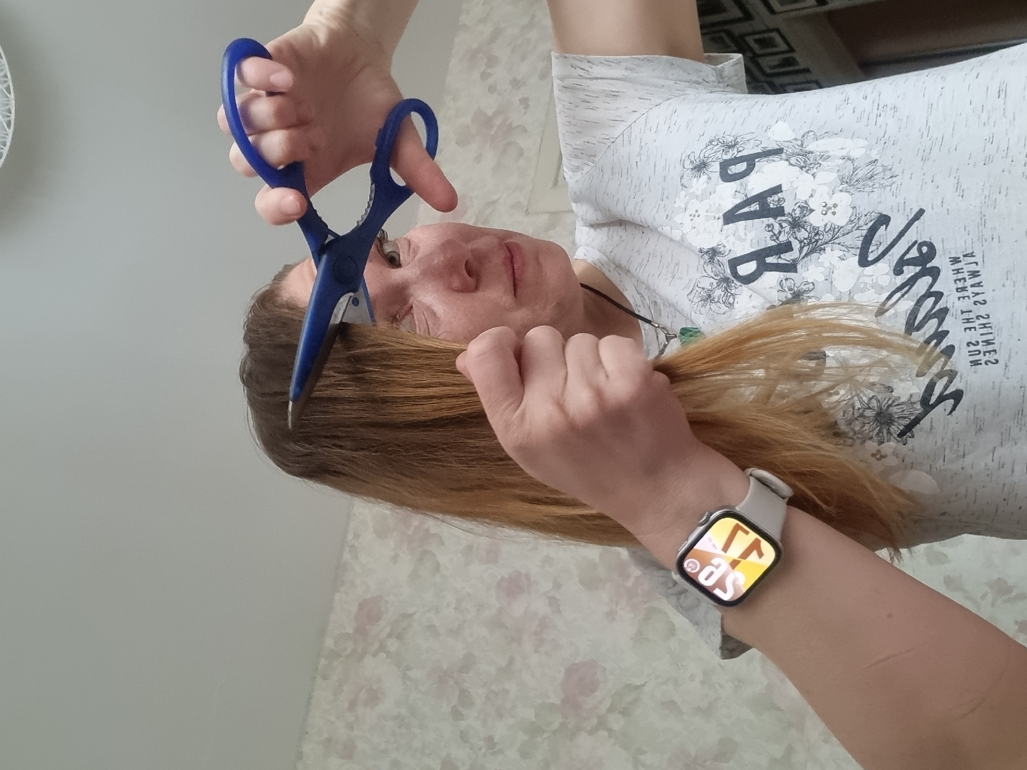 An individual, likely female, with light brown hair, is depicted indoors, attempting to cut their own hair. The subject, viewed from the chest up, holds a section of hair with their left hand in front of their right shoulder, while their right hand operates a pair of blue-handled scissors positioned to cut the hair. A silver-cased smartwatch with a white band is visible on the left wrist, displaying a bright, multi-colored interface. The subject wears a light grey t-shirt featuring a mirrored floral design and text, and a dark necklace with a green pendant. The background consists of a light-colored wall with a faint floral wallpaper pattern on the left, and a plain white ceiling. The scene is located in Chaikovsky, Russia.