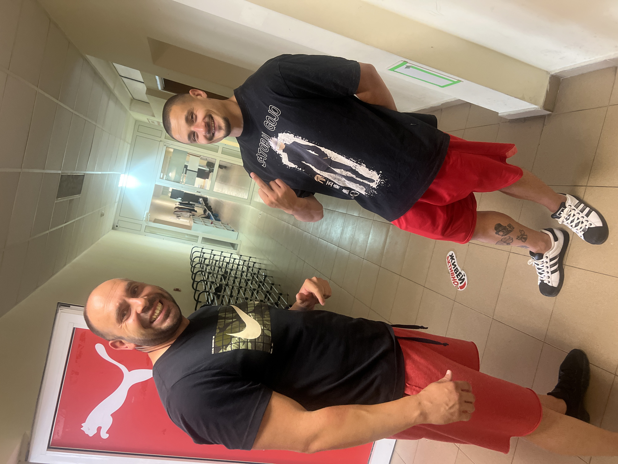 Two men are posing in a hallway. One man is in the foreground next to a red Puma sign. He is wearing a black t-shirt with a white Nike logo, red shorts, and black shoes. He is bald and smiling. The other man is in the background wearing a black t-shirt with a white graphic, red shorts, and white and black Adidas sneakers. He is also smiling and pointing with his left hand. There is a sticker on the floor that reads "Живеем Активно". A coat rack sits against the wall in the background. The location is the city of Sofia, Bulgaria.