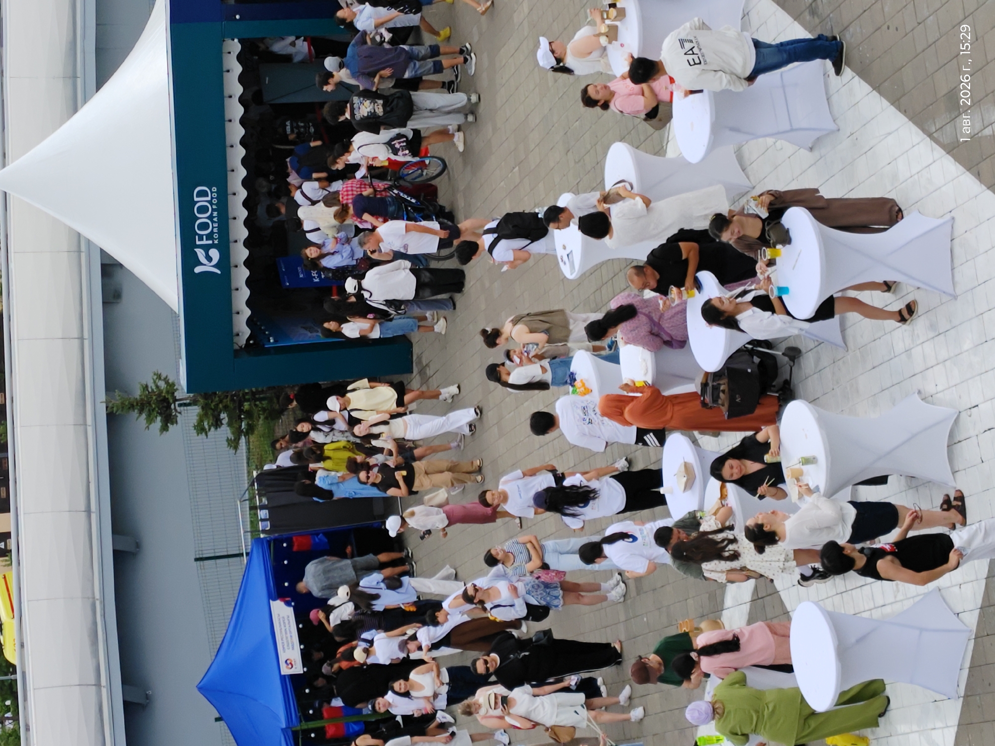 The image depicts an outdoor food festival with a Korean food theme, identified by the "K FOOD KOREAN FOOD" signage on a white tent. The event appears to be taking place on a paved plaza in Astana, Kazakhstan, as indicated by the timestamp "1 авг. 2026 г., 15:29" which translates to August 1, 2026, 15:29.
A diverse crowd of adults, primarily young and middle-aged, are milling about the area. They are dressed in casual summer attire, including t-shirts, shorts, skirts, and light jackets. Many are standing in small groups, some interacting at white, high-top tables set up for food and drinks. Others are walking, queuing, or observing the activities.
Several stalls or booths are visible, including a blue tent on the left and the prominent white tent housing the Korean food vendor. Some people are seen interacting with the stalls, making purchases, or receiving food. Food and drinks are visible on the tables, including cups and what appear to be take-out containers.
The environment is bright and appears to be daytime, with a clear sky. The ground is paved with square tiles. In the background, a portion of a large building with a gray facade is visible, along with a fence and some greenery. The overall atmosphere suggests a lively and social event.
