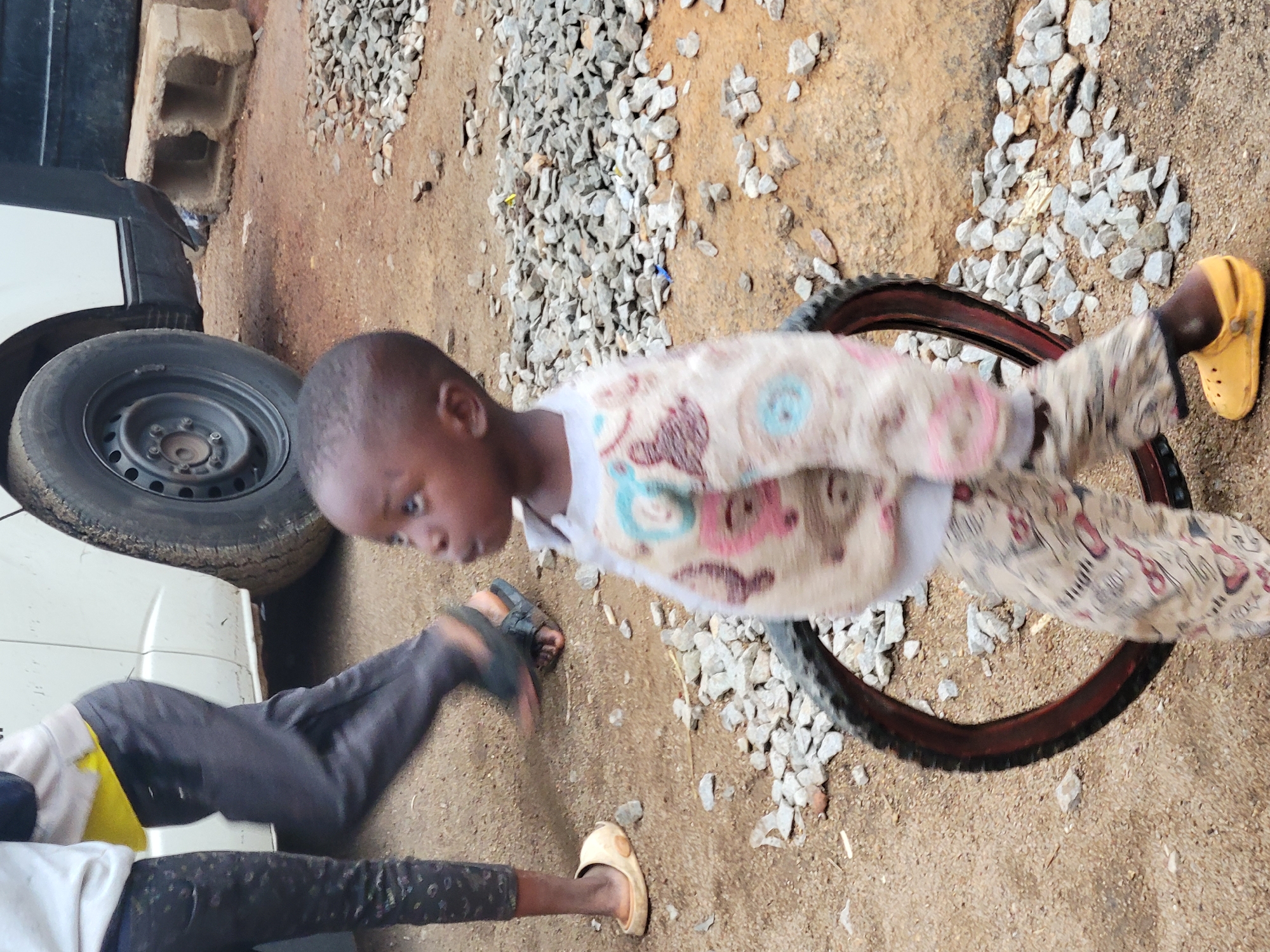 The scene is an outdoor area in Jos, Nigeria. The ground is a mixture of dirt and small rocks. A young Black child is standing inside a tire rim. The child is wearing patterned pajamas and yellow sandals. Nearby, another person's lower body is visible. They are wearing dark pants and light-colored sandals. A white vehicle with a black tire is visible on the left. A concrete block rests next to the vehicle.