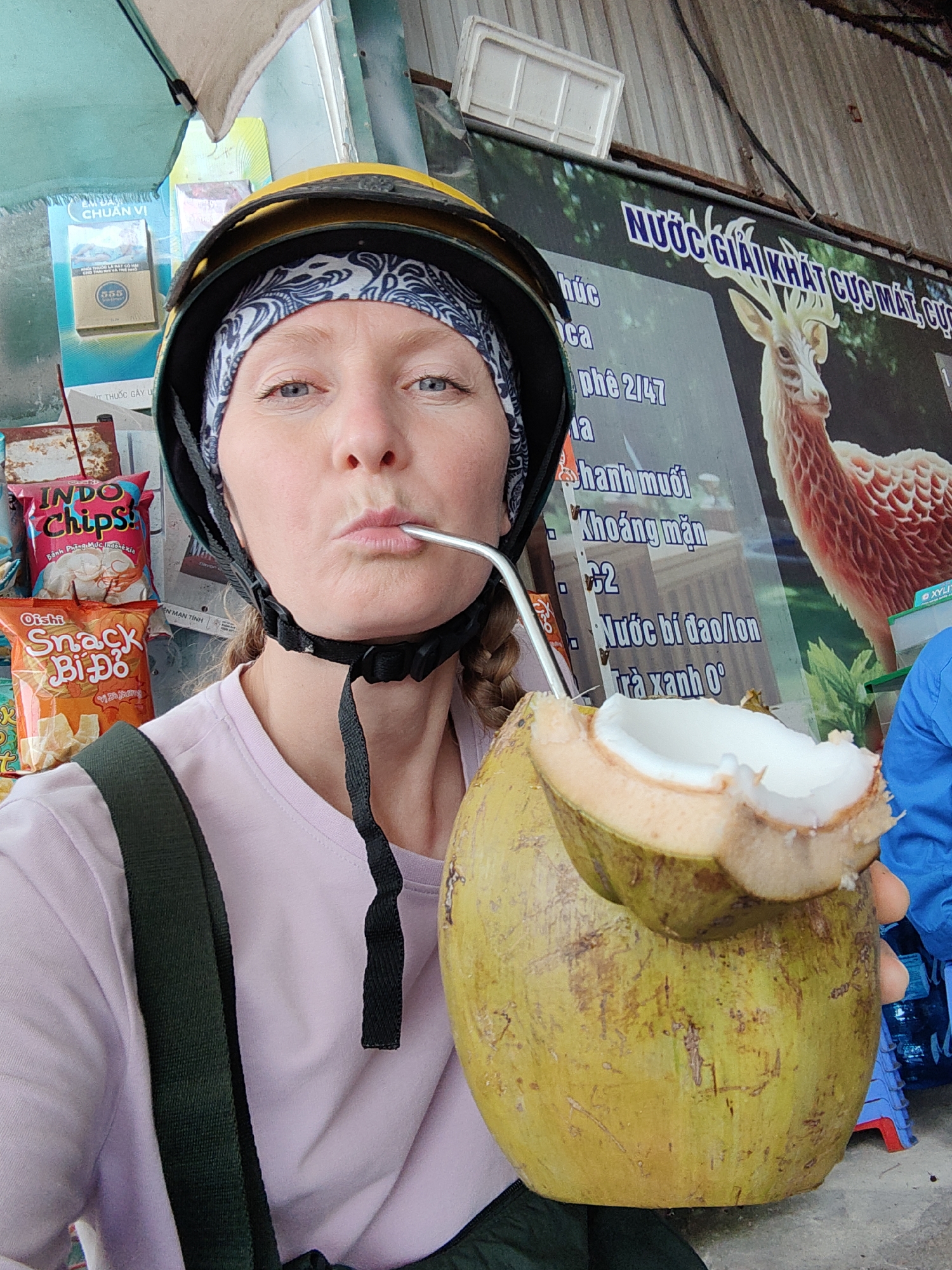 A woman wearing a yellow motorcycle helmet and a black and white headscarf is drinking from a coconut with a metal straw. She has fair skin, blue eyes, and her hair is braided. She is wearing a lavender t-shirt and a dark green backpack strap is visible on her left shoulder.
In the background, there are snack foods, including "Indo Chips" and "Oishi Snack Bi Do," as well as a sign with the words "NƯỚC GIẢI KHÁT CỰC MÁT, CỰC" written on it, along with an image of a deer. Other Vietnamese text is also visible. The environment appears to be an outdoor vendor stand. The location is likely Cao Xanh Ward, Vietnam.