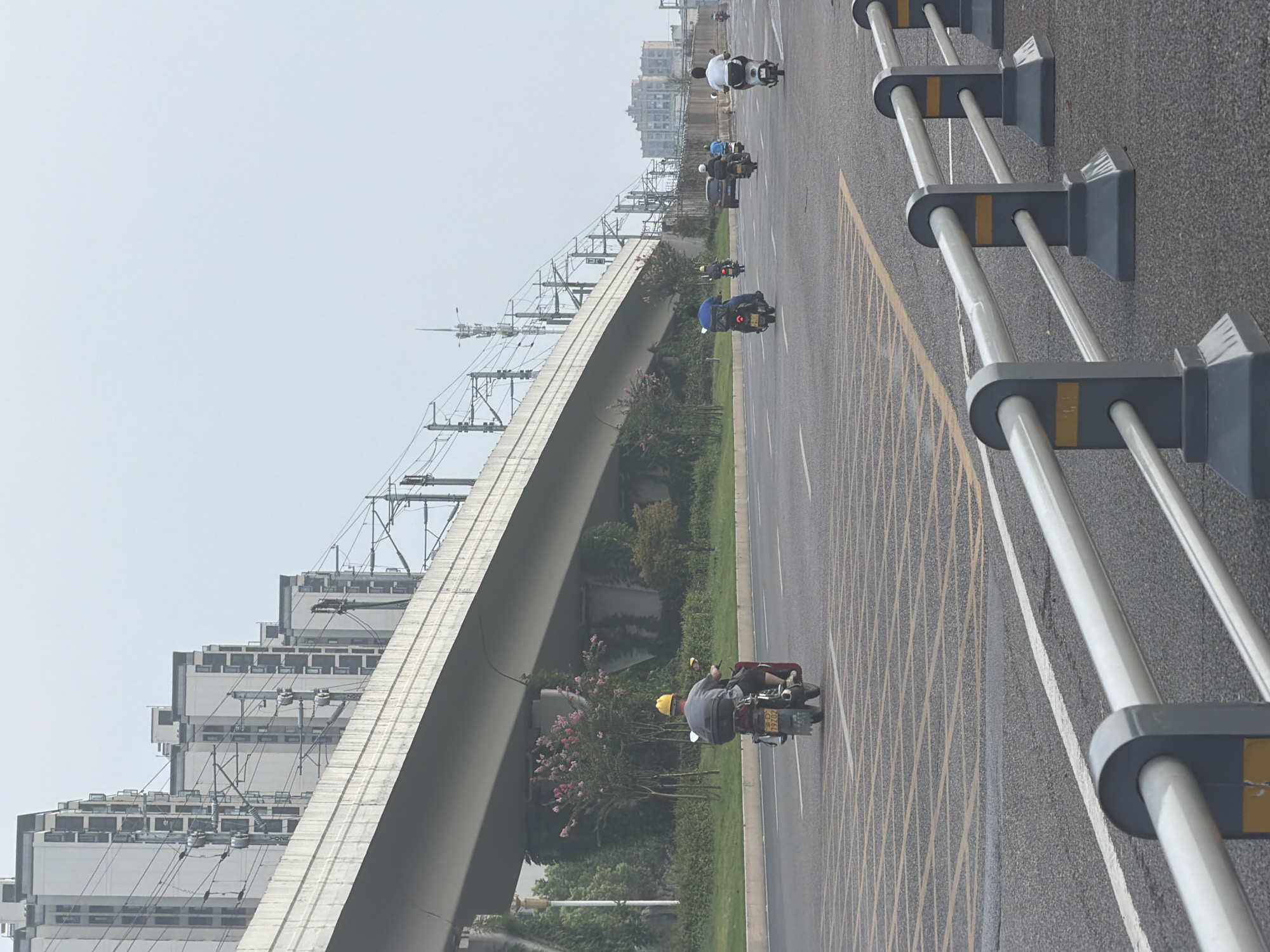 The image depicts a street scene in Jiangjin District, Chongqing, China, under an elevated transportation structure, likely an overpass for a railway or highway. The sky is overcast. In the foreground, a silver metal barrier with gray posts and yellow accents runs along the side of the road. The road itself has yellow grid markings and white lane dividers. Several individuals are riding motorbikes and scooters on the road, moving in the same direction. One rider in the foreground wears a yellow helmet. In the background, a row of modern, multi-story apartment buildings are visible, aligned with the elevated structure. Greenery, including trees and bushes, lines the side of the road beneath the overpass. Further in the distance, more buildings and infrastructure can be observed.