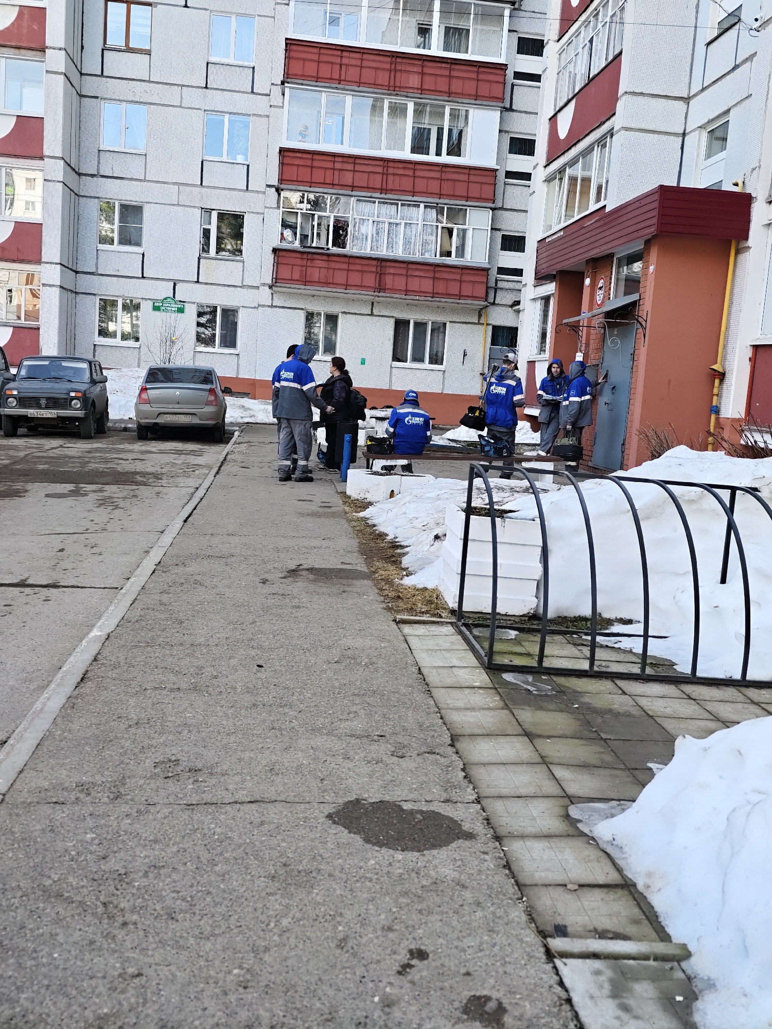 The image shows an outdoor scene in the city of Chaikovsky, Russia.
Several people wearing blue Gazprom work uniforms are gathered near the entrance of an apartment building. One person not in uniform appears to be speaking with them.
Two cars are parked nearby; a dark-colored Lada Niva and a gray Renault Logan.
The architecture is characterized by a multi-story residential building with a mix of white and red exterior finishes. The sidewalk and ground have patches of snow, indicating a cold or winter season.
There is a metal structure near the sidewalk covered with snow, that appears to be some sort of planter or garden enclosure.