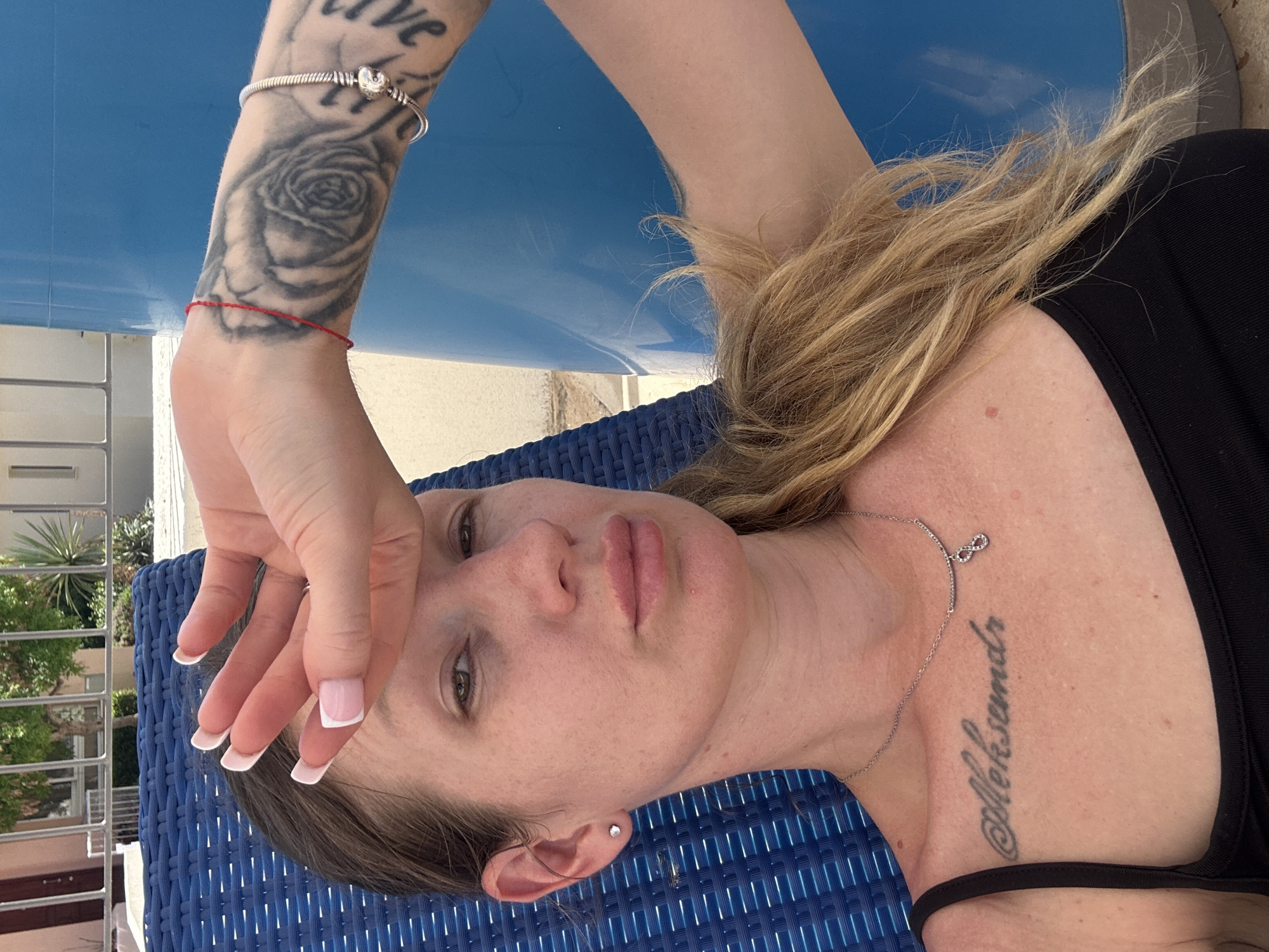 A woman with blonde hair pulled back in a ponytail is lying on a blue lounge chair. She is wearing a black tank top and a silver necklace with an infinity charm. She has a tattoo on her chest that reads "Aleksandr" and another tattoo on her arm that appears to be a rose. Her left hand is raised to her forehead. She is wearing a small diamond stud earring in her left ear. A thin red string bracelet is on her right wrist. She is outdoors with a blue cylindrical object in the background, possibly part of a swimming pool. The environment suggests a sunny location, likely a resort or hotel in Peyia, Cyprus.