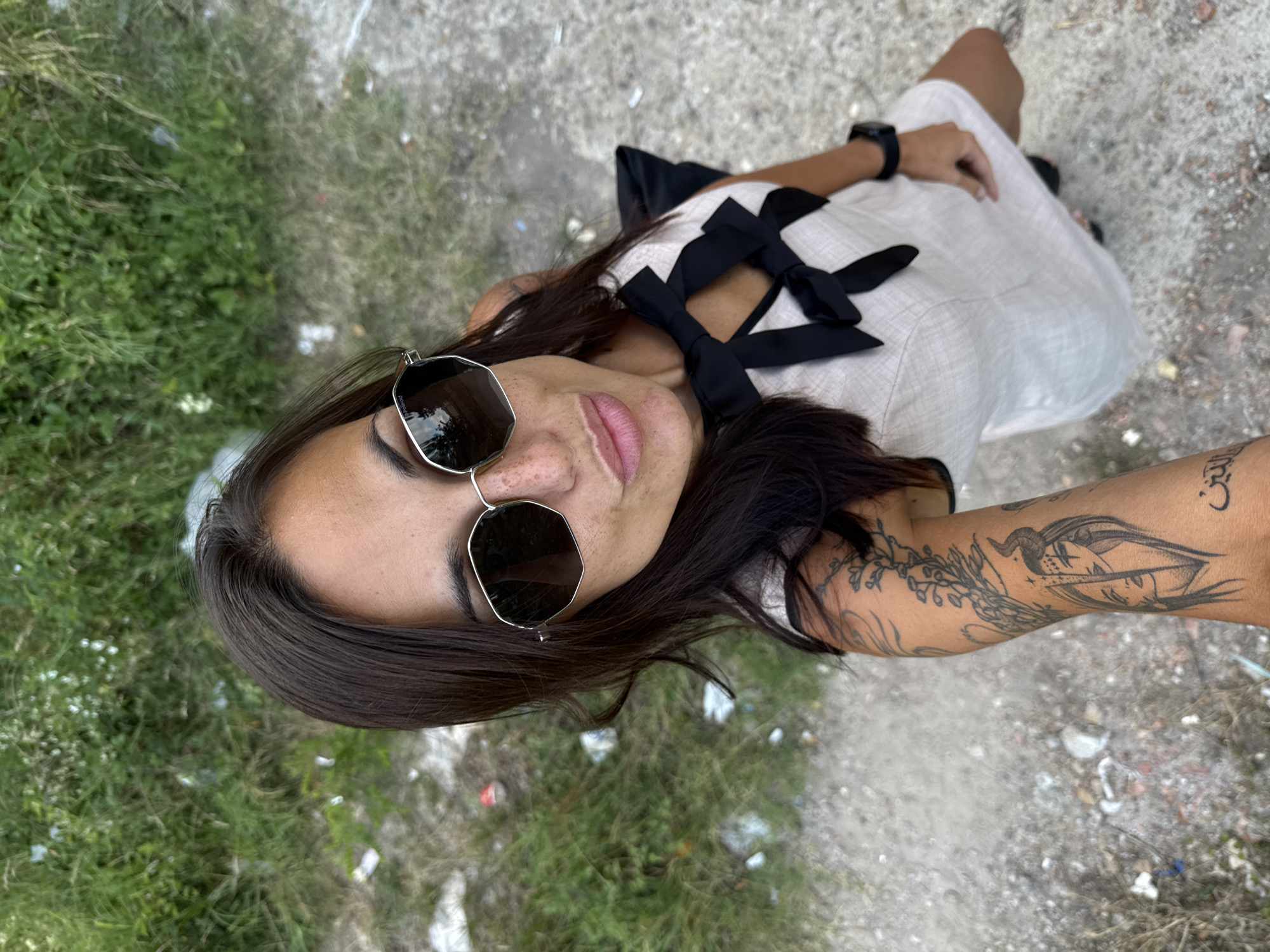 In the city of Pleven, Bulgaria, a woman is captured from a high-angle perspective, holding a camera to take a selfie. She is wearing a light-colored dress with black bows on the chest and sunglasses. Her left arm, adorned with tattoos, is visible. The background consists of green vegetation and a gravelly ground, with scattered debris indicating a potentially neglected area.