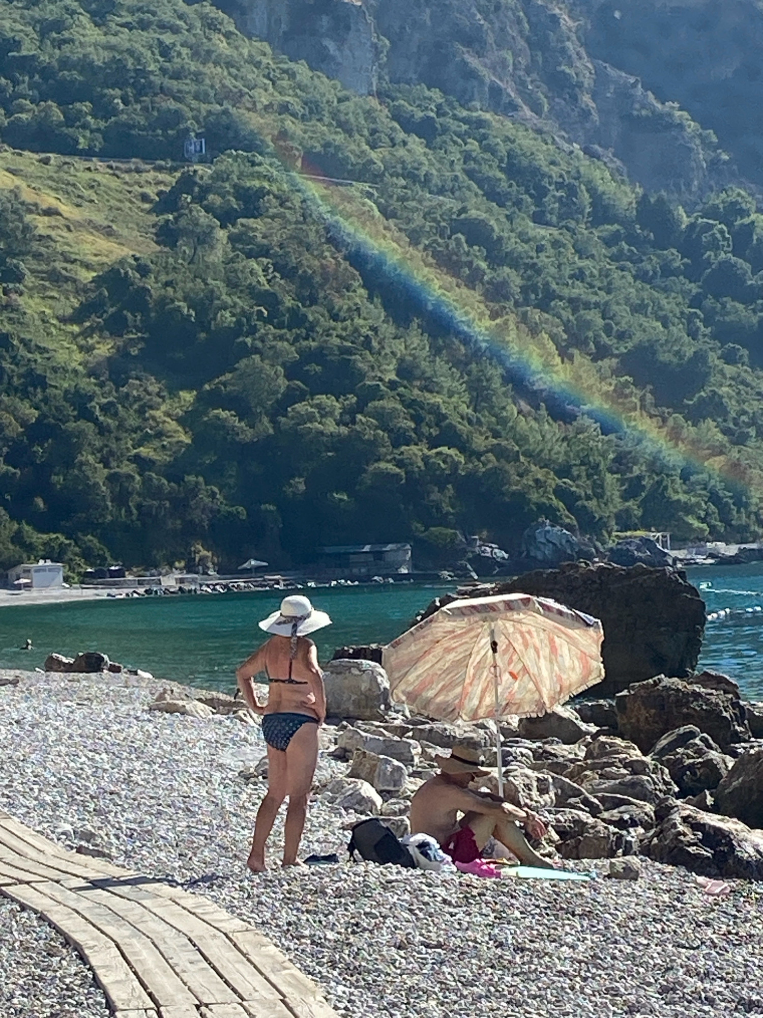 This image captures a coastal scene near Prijevor, Montenegro. In the foreground, a wooden walkway leads towards a pebble beach. Two individuals are present on the beach. One person, wearing a wide-brimmed hat and a bikini, stands facing away from the camera. Another person, wearing a straw hat and red shorts, is seated under a striped umbrella, facing left. The background features the turquoise water of the Adriatic Sea, a rocky coastline, and a lush, tree-covered mountainside where a faint rainbow is visible.