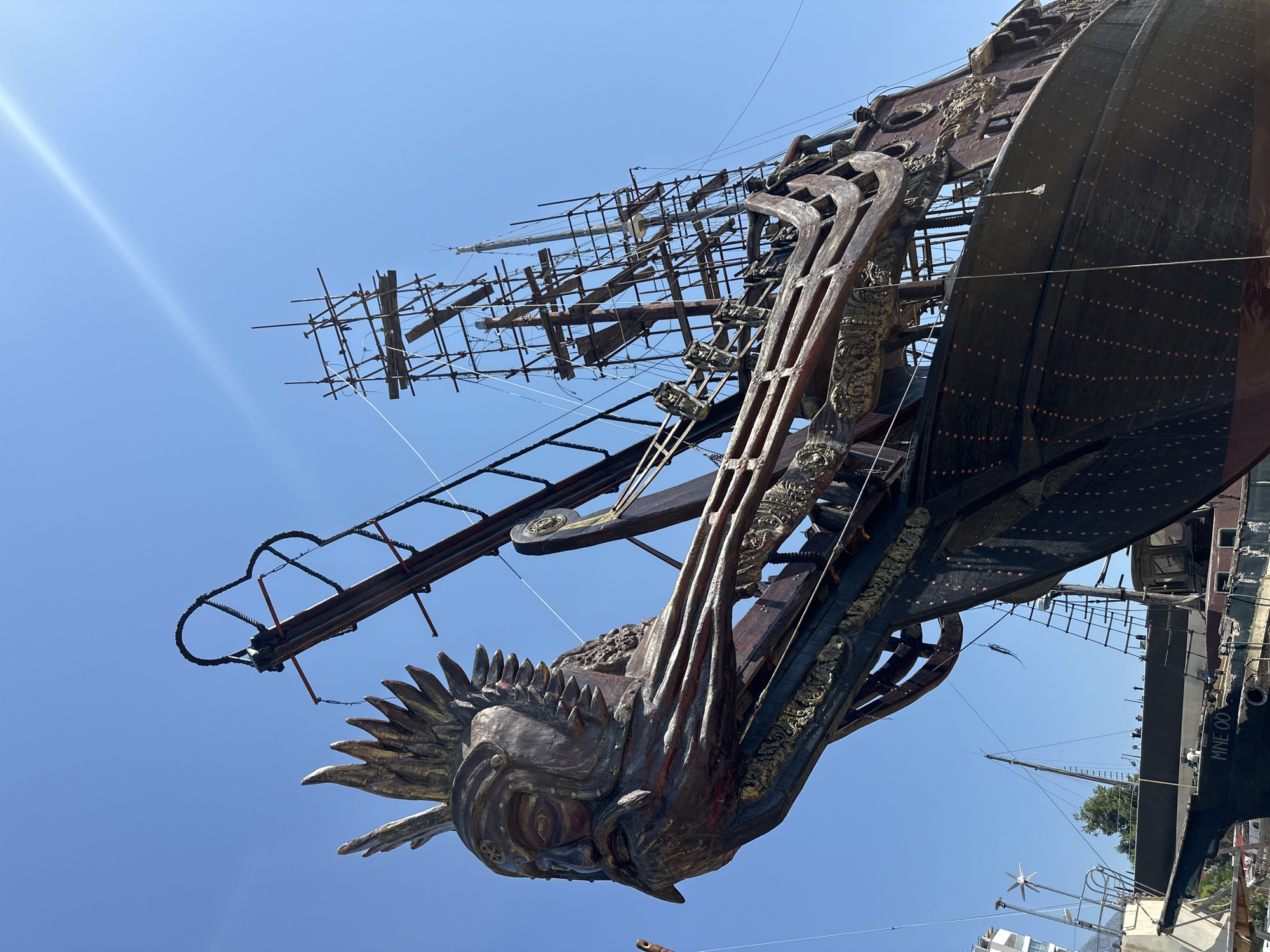 The image shows the bow of a wooden sailing ship, likely a replica or prop, adorned with a large, ornate figurehead of a human face with a headdress. The ship is dark brown and appears to be in a dry dock or shipyard, with scaffolding surrounding its masts and rigging. The sky is a clear, bright blue. In the background, other vessels and parts of shipyard infrastructure are visible. The overall impression is of a construction or restoration project for a historical or fantasy-themed ship. The ship is located in the city of Bar, Montenegro, specifically in the shipyard area.
