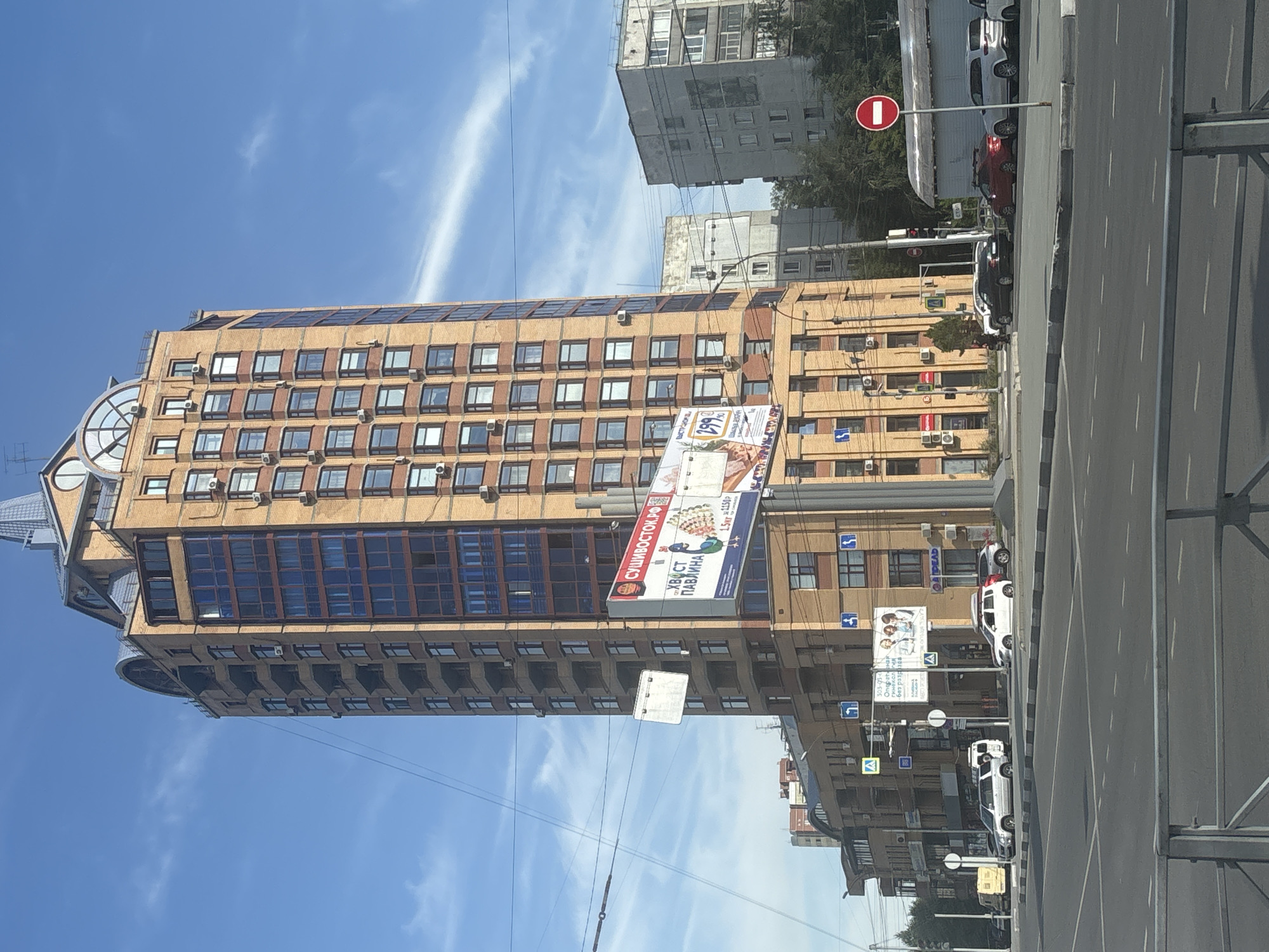 The image captures a sunlit urban street scene in Novosibirsk, Russia, dominated by a tall, multi-story brick building with numerous windows and balconies, featuring a distinctive curved roofline. In the foreground, a road with lane markings and a pedestrian crossing is visible. Several cars are parked along the roadside and driving on the street. A large billboard advertising "Sushivostok.ru" with an image of a peacock and text in Russian is prominently displayed on a pole. To the right of the main building, a lighter-colored, less ornate apartment building stands. The sky is clear blue with scattered white clouds. There are no discernible people actively engaged in interactions. The scene appears to be a typical daytime view of a city intersection with commercial and residential structures.