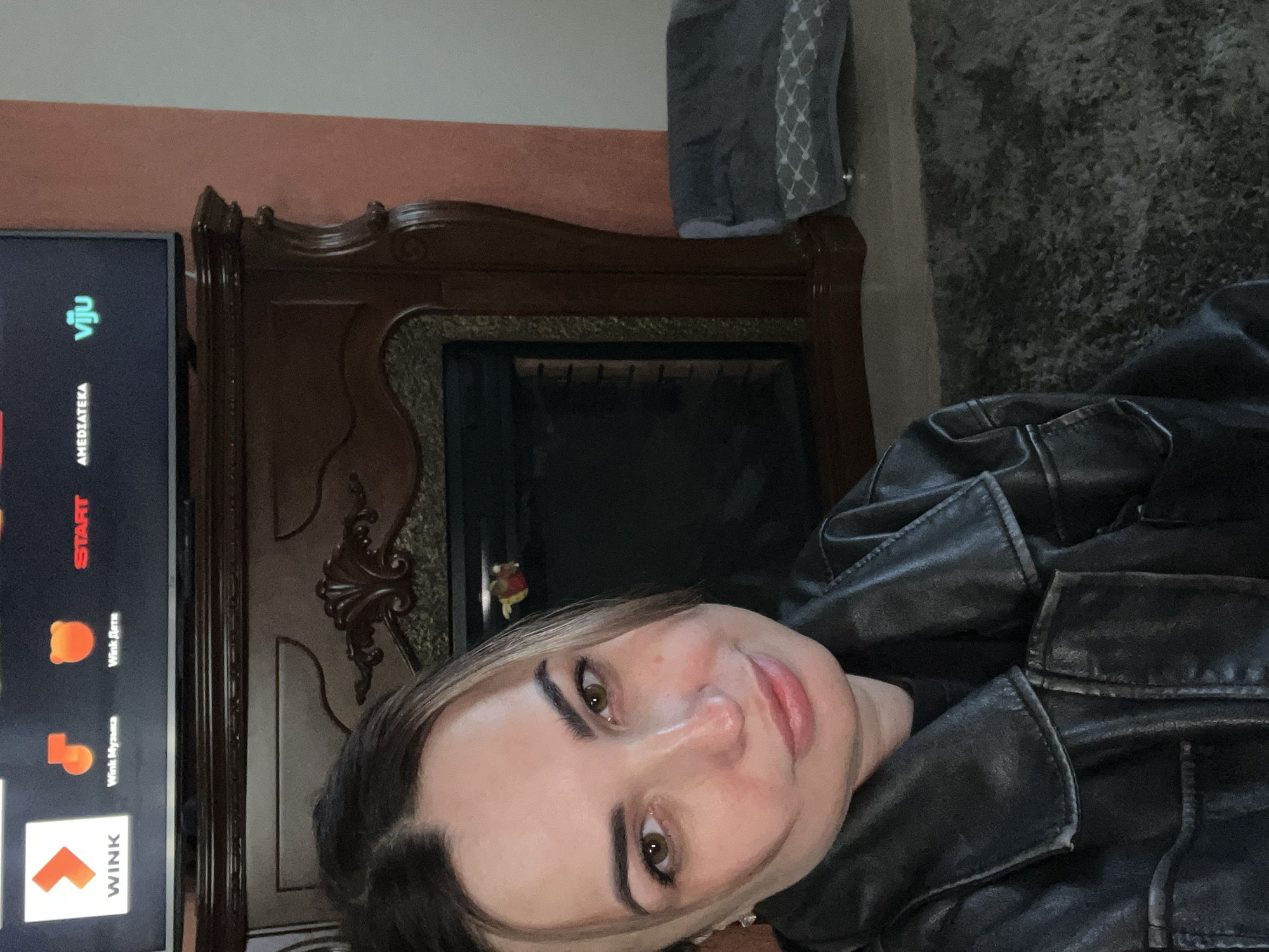 A selfie shows a woman with fair skin, light brown hair, and brown eyes, wearing a black leather jacket. She is positioned in front of a dark brown fireplace with intricate wood detailing. To the left of the fireplace is a flat-screen TV displaying app icons, including "WINK" and "VIJU". The background behind the fireplace is a peachy-orange wall, and to the right of the fireplace, there appears to be a gray throw or cushion. The location and city are unknown.