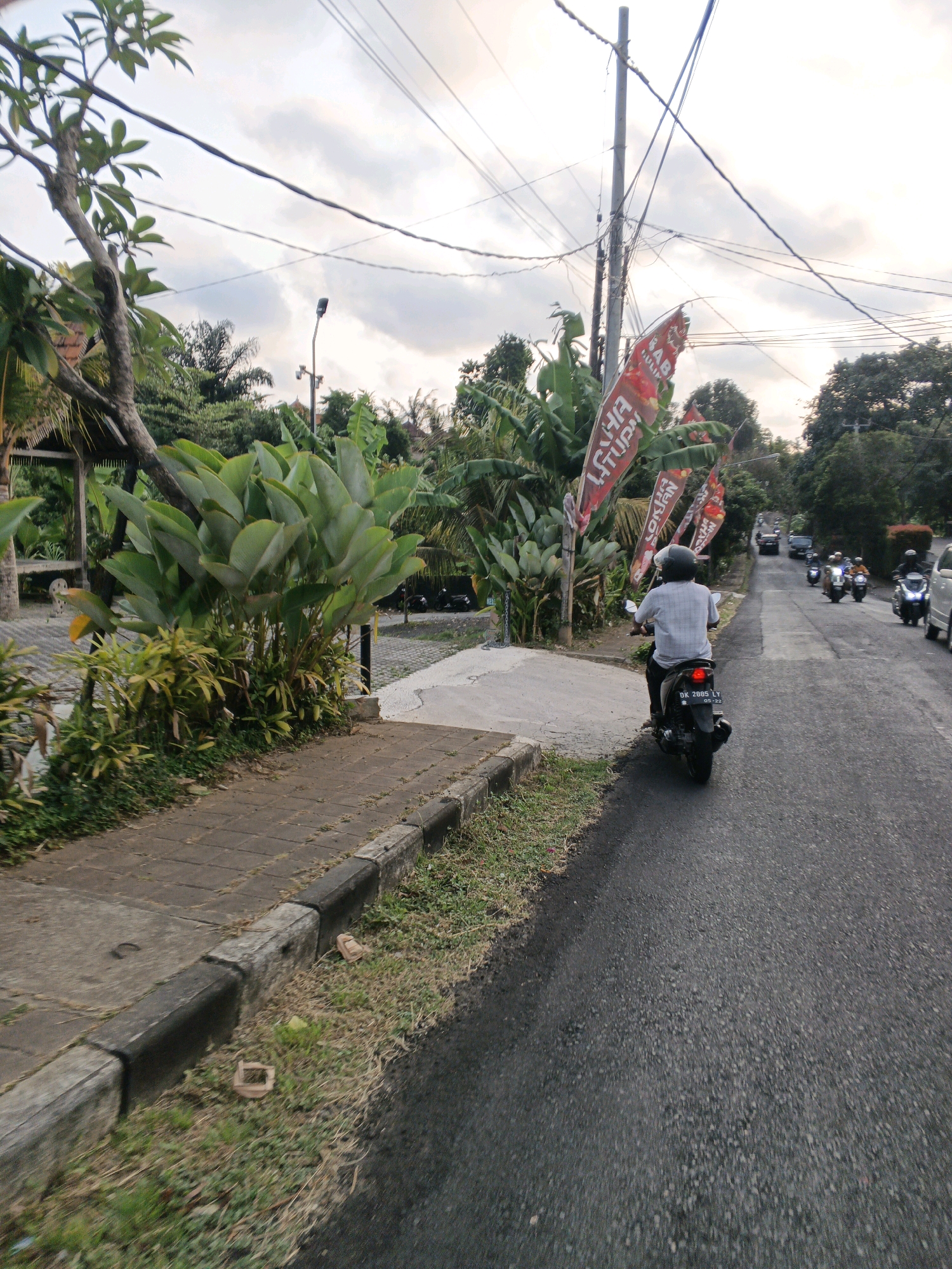 The image depicts a street scene in Sukawati, Indonesia. A person is riding a motorcycle on a paved road. The motorcycle is black with a license plate that reads "DK 2085 LY 05-22". The rider is wearing a white shirt and a black helmet. Red flags with white writing are planted along the side of the road. Large green plants are visible on the left side of the image. The sky is overcast with clouds. In the background, other motorcycles and vehicles are seen on the road. The road is lined with a sidewalk made of interlocking pavers. The environment appears to be tropical.