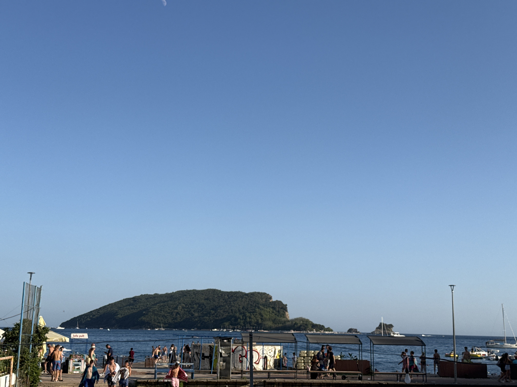 The image shows a clear, blue sky with a small crescent moon visible in the upper left corner. Below the sky, a body of water, likely the Adriatic Sea, stretches across the frame. On the water, several boats and a large, green, tree-covered island are visible in the distance. In the foreground, a pier or promenade runs along the waterfront. A number of people, dressed in casual summer clothing, are walking along the pier. Some appear to be returning from the water, carrying towels. There are also some covered seating areas and structures along the pier. The scene suggests a warm, sunny day in a coastal city, likely Budva, Montenegro, known for its scenic coastline and proximity to islands.