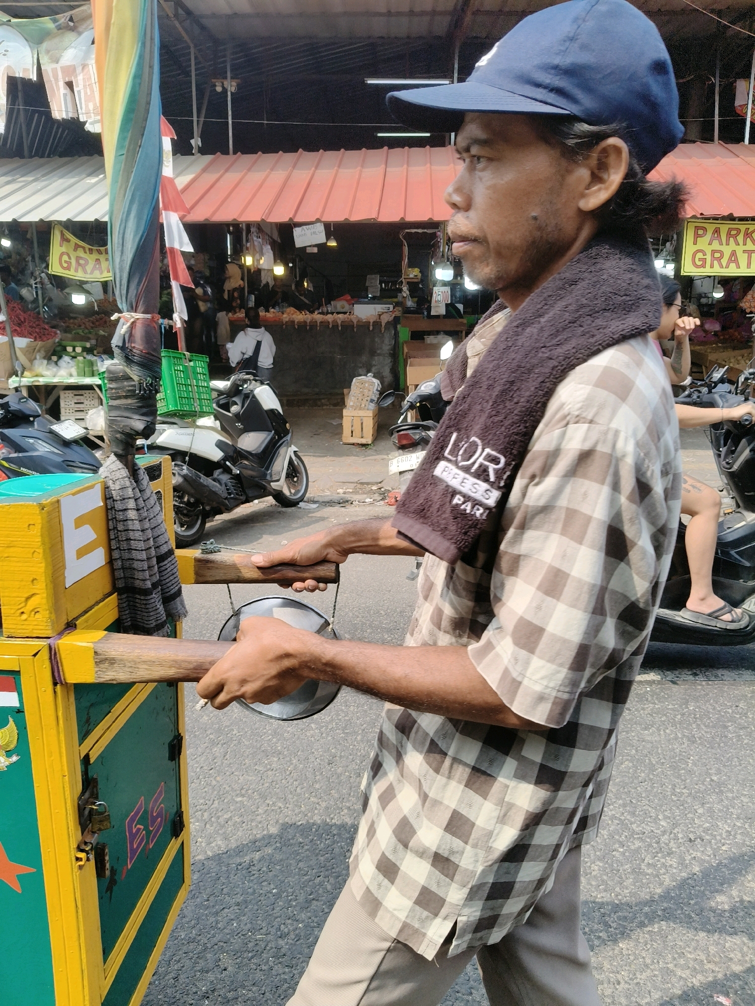 This is a medium shot photograph taken from the side of a man pushing a food cart in an outdoor market. The man is wearing a blue baseball cap, a brown and white plaid shirt, and beige pants. A dark brown towel with embroidered white lettering is draped around his neck. He is holding onto the handles of the food cart, which is yellow and green with some red and white decorative elements. The cart has various containers and what appears to be a metal basin attached to it.
In the background, there are market stalls with awnings, displaying various goods. There are also several motorcycles parked in the vicinity. One stall has a sign with yellow lettering that reads "PARK GRATIS." The ground is paved with asphalt. The overall scene suggests a busy street market environment. Based on visual cues, this scene is likely located in an urban setting in South Tangerang, Indonesia, a region known for its bustling markets and street vendors.