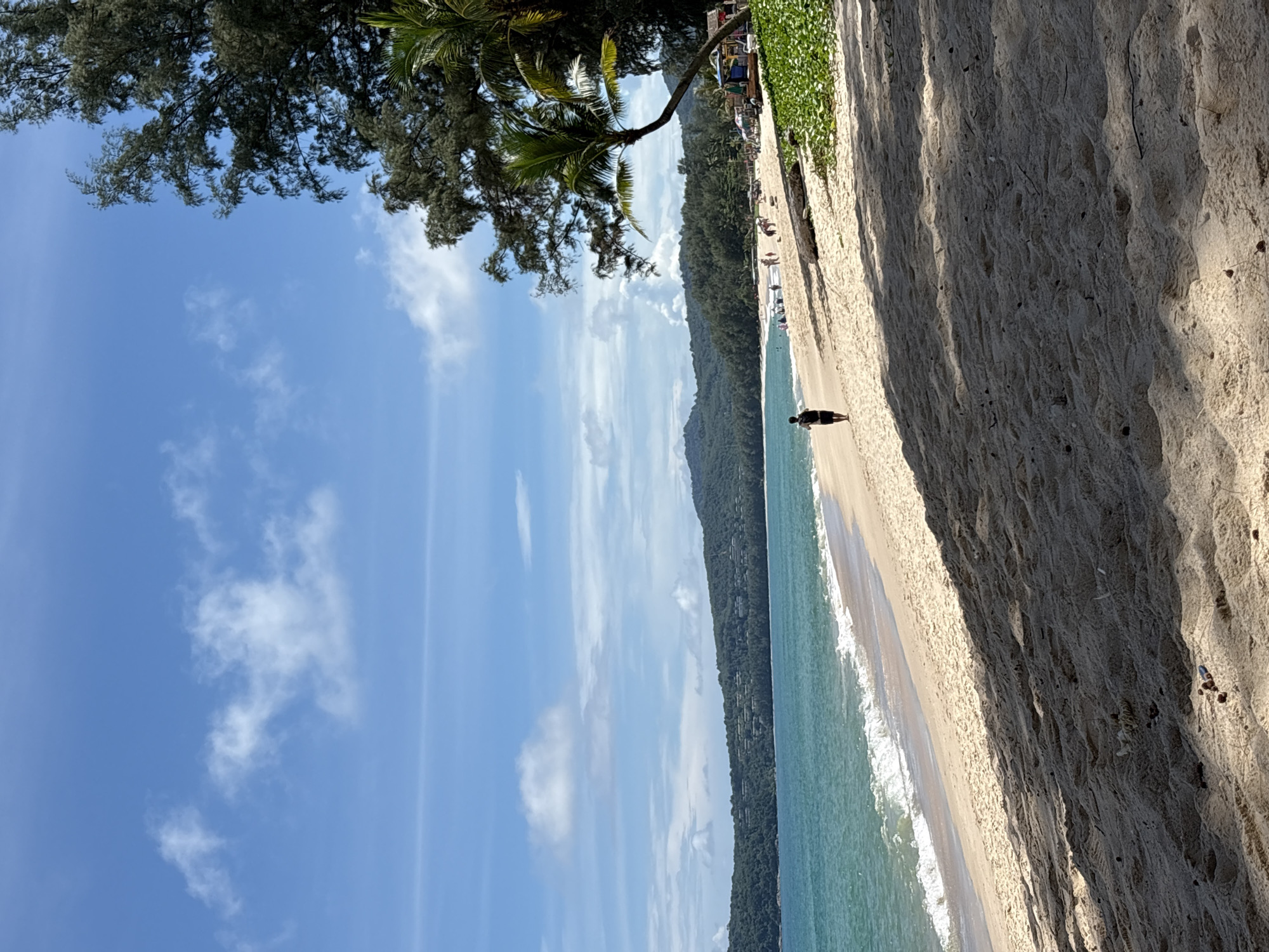 This is a photograph of a beach in Choeng Thale, Thailand. A single person is walking away from the camera on the wet sand near the water's edge. The ocean is a vibrant turquoise and gentle waves are lapping at the shore. In the background, lush green hills covered in trees form a backdrop to the beach, with a few buildings nestled among the foliage. The sky is a bright blue with scattered white clouds. To the right of the frame, several palm trees are visible, their fronds casting shadows on the sand, and a small cluster of beach umbrellas and seating areas are present. The sand in the foreground is textured with footprints and scattered debris.
