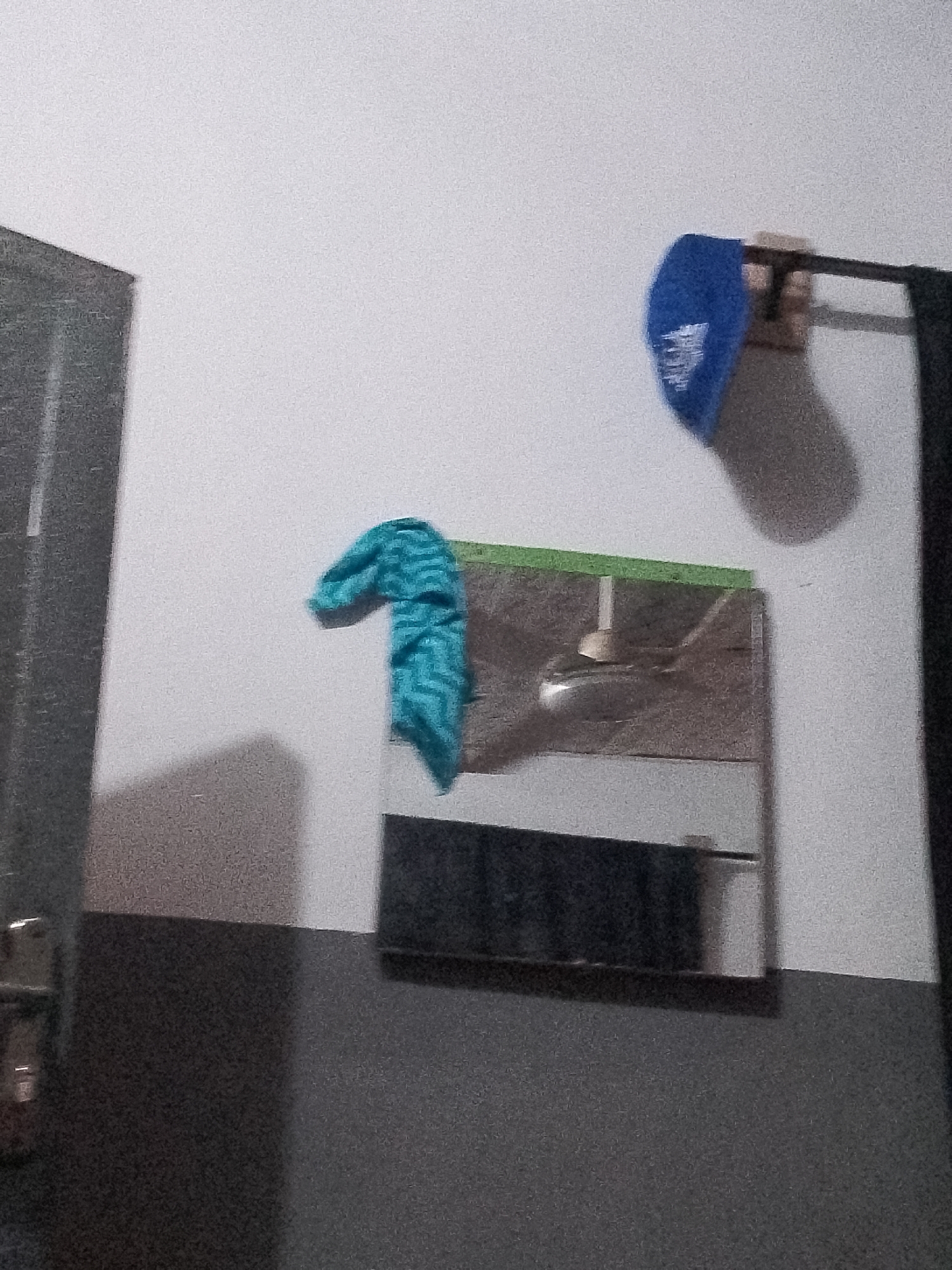 A blue baseball cap with white lettering is hanging from a hook on a white wall. To the left, a blue and white striped towel is draped over a mirror. The mirror is mounted on a green-taped surface and reflects a ceiling fan. A dark gray door frame with a silver handle is visible on the left side of the image. The lower part of the wall is a dark gray color. The scene is set in an indoor environment in Jauro Yinu, Nigeria.