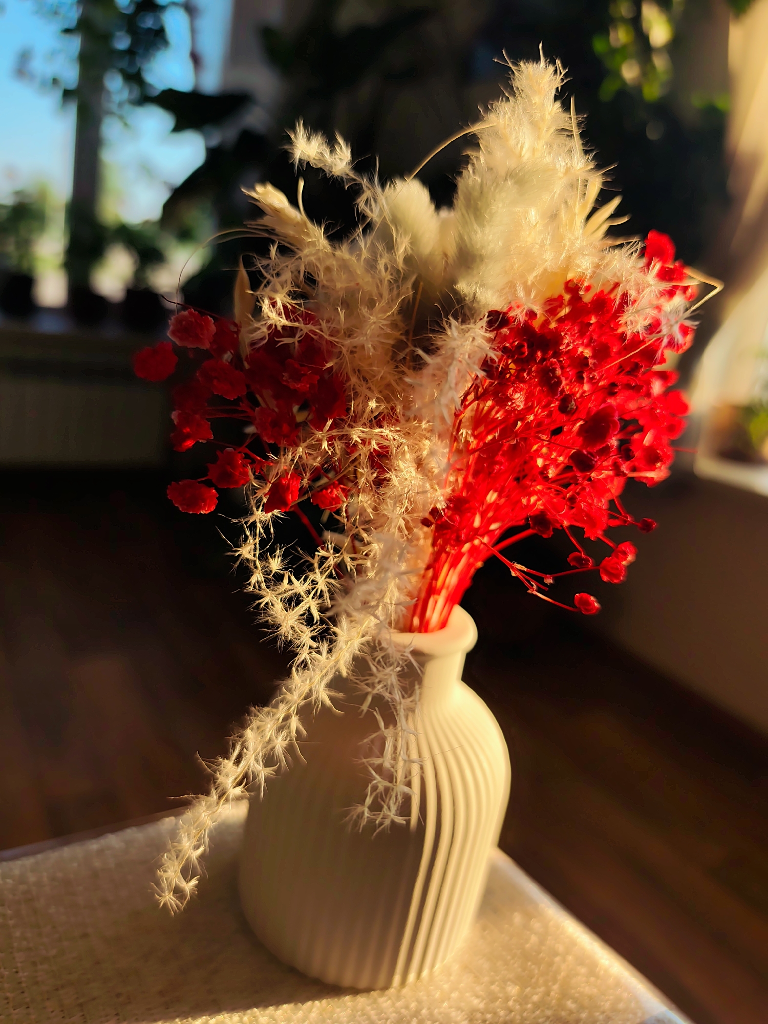 A vase containing dried red and white flowers sits on a table. The vase is light-colored and vertically ribbed. The dried flowers are a mix of red and feathery white stems. The background is blurred, showing a window with plants visible outside. The light suggests it might be late afternoon or early morning. The location, Нижний Пальник, Russia, is not visually verifiable from the image.