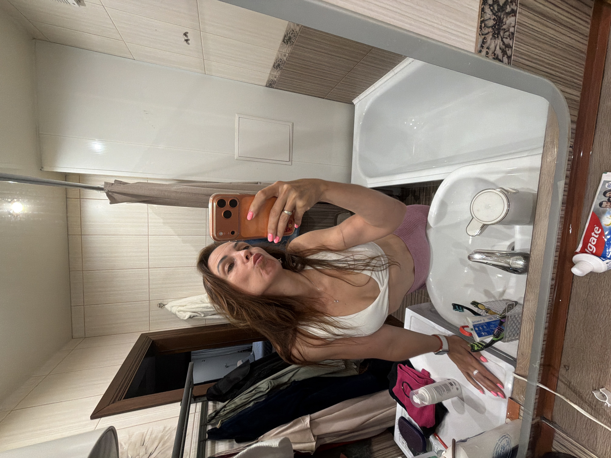 A woman is taking a mirror selfie in a bathroom. She is wearing a white sports bra and pink shorts. She has long brown hair and is wearing nail polish. The bathroom has tiled walls and a white bathtub. There is a sink with toiletries on the counter. Clothing is hanging on a rack behind the woman. A tube of Colgate toothpaste is visible in the foreground. The location is specified as Birobidzhan, Russia, although this detail cannot be visually confirmed from the image.