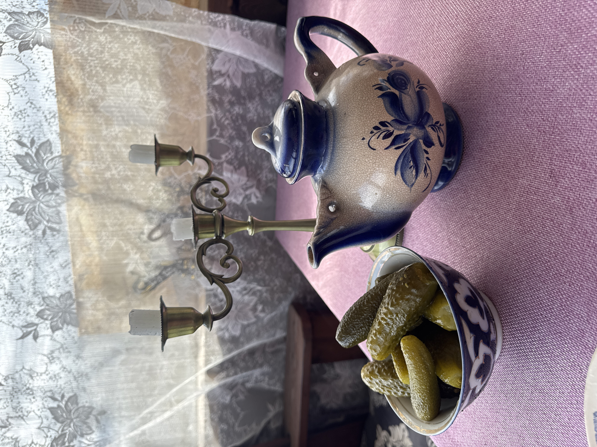 The image shows a still life arrangement. In the foreground, a bowl filled with pickled cucumbers is visible. Adjacent to the bowl, there is a blue and beige ceramic teapot. Behind these objects, a brass candelabra with three white candles stands. A white, floral-patterned curtain serves as a backdrop. The scene is set on a lilac-colored surface, likely a tablecloth. The exact location in Chaikovsky, Russia, cannot be determined from this image.