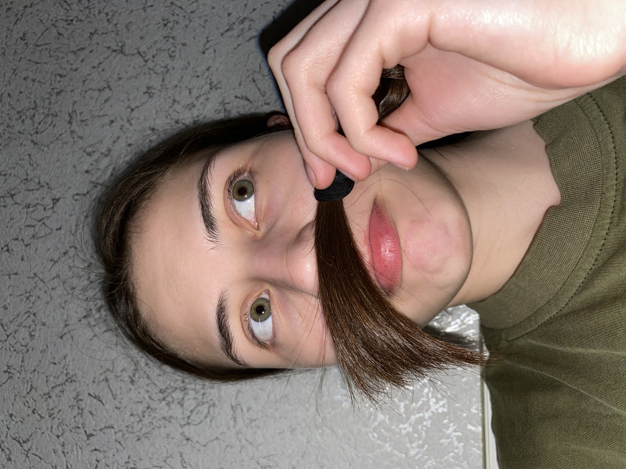 A young woman is oriented such that her face is in the center, tilted slightly towards the viewer's left. She has fair skin, light brown hair, and green eyes. A section of her hair is pulled down and held under her nose with a black hair tie, resembling a mustache. Her lips are pink. She is wearing an olive green shirt. The background appears to be a textured gray wall. There is no indication of the location being Votkinsk, Russia, or any other city.