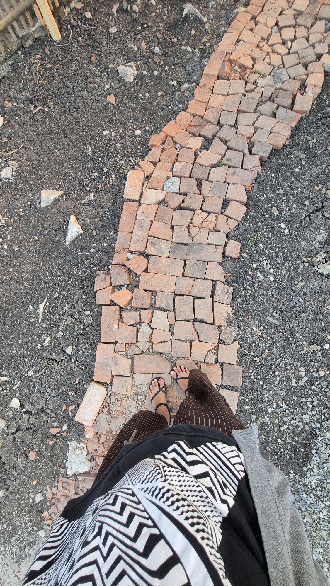In Gambiranom, Indonesia, an overhead shot captures a person standing on a path made of broken, irregularly shaped red bricks. The path is narrow and curves slightly to the left, bordered by dark, dry earth and gravel. The person is wearing black flip-flops, brown pinstriped pants, and a black shirt with a bold black and white zigzag pattern, partially covered by a grey cardigan. The environment appears to be an outdoor, undeveloped area, possibly a construction site or a neglected pathway, with scattered debris and small green shoots growing between the bricks.