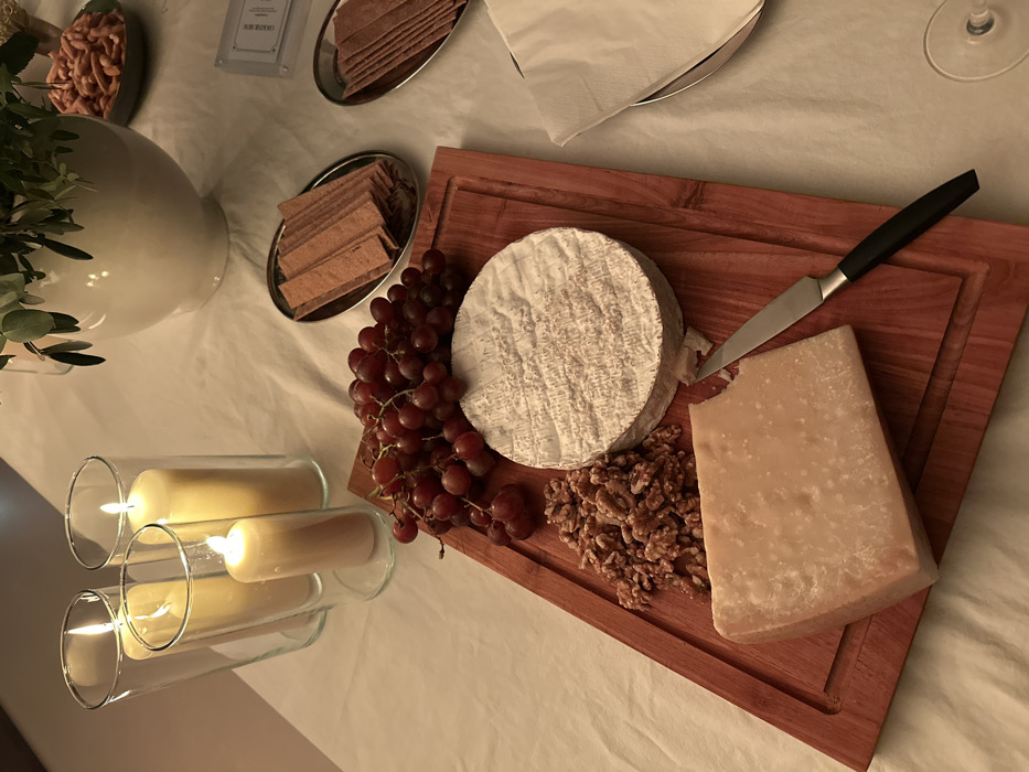 The image displays an indoor scene focused on a table set with food and ambient lighting. A white tablecloth covers the table surface. On the left, four lit pillar candles are enclosed within clear glass hurricane lamps of varying heights, emitting a warm glow. A central reddish-brown wooden cutting board presents a large cluster of red grapes, a round wheel of soft white-rinded cheese, a pile of shelled walnuts, and a block of firm, light-yellow cheese, with a small portion appearing cut. A small cheese knife with a silver blade and black handle rests next to the firm cheese. To the right, two oval silver-colored trays each hold stacks of reddish-brown rectangular crackers. Further back, a white ceramic vase contains green foliage, and a small dish holds numerous orange-colored snacks. A folded white napkin on a silver plate and the base of a wine glass are visible in the lower right. No individuals are present, and no specific actions or interactions are depicted. The precise location within the city of London, United Kingdom, cannot be determined from the visual information provided.