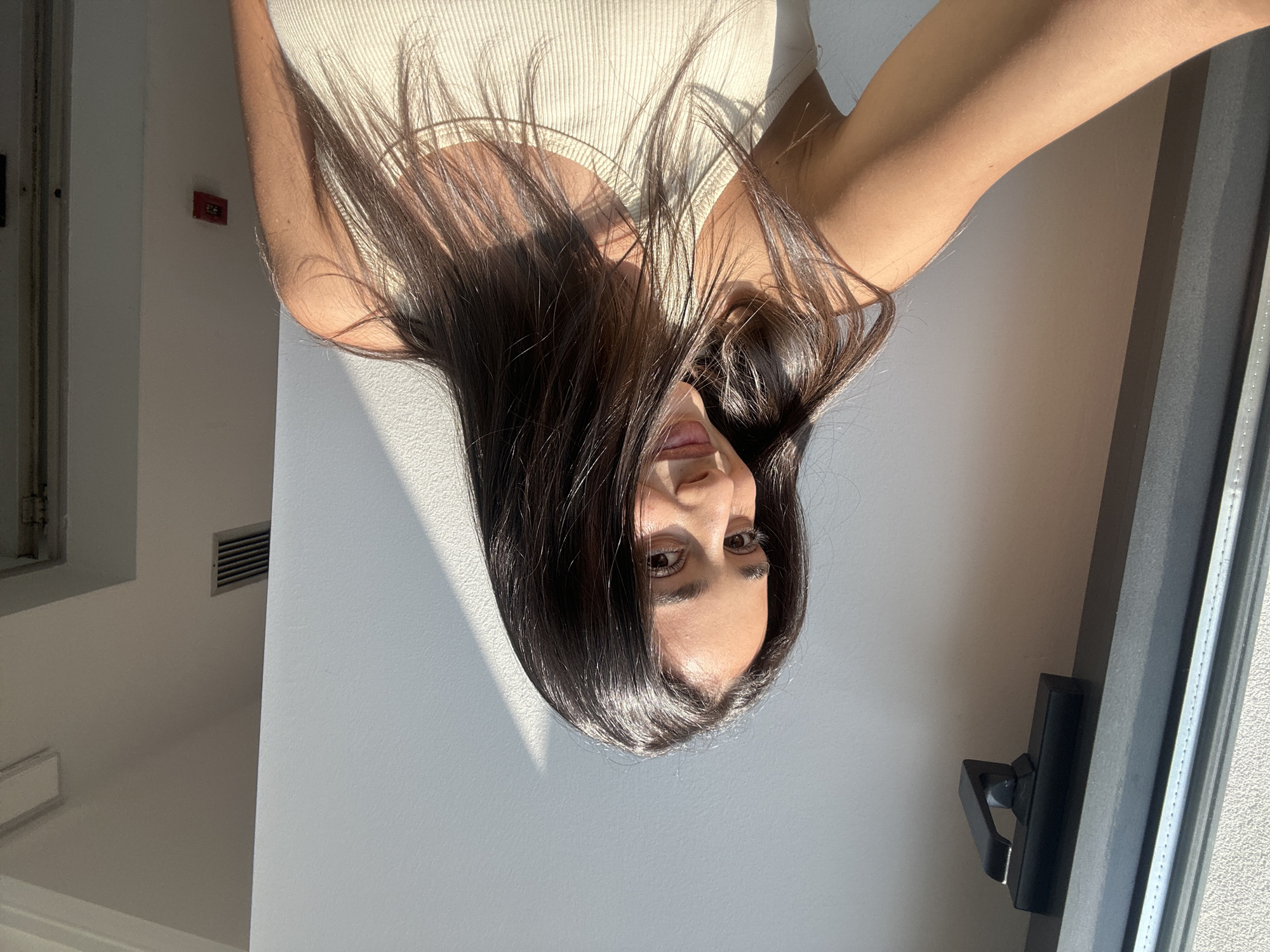 A young woman with long, dark hair is illuminated by sunlight streaming through a window on the left side of the frame. She is looking towards the camera with a slight smile, her face partially obscured by her hair. She is wearing a light-colored, ribbed tank top. The background is a plain white wall, with a shadow cast by the window frame diagonally across it. A dark grey window frame is visible on the far left. A red fire alarm is mounted on the wall in the background on the right. The scene appears to be indoors, possibly an apartment or hotel room, with no discernible urban context that would specifically place it in Milan, Italy.