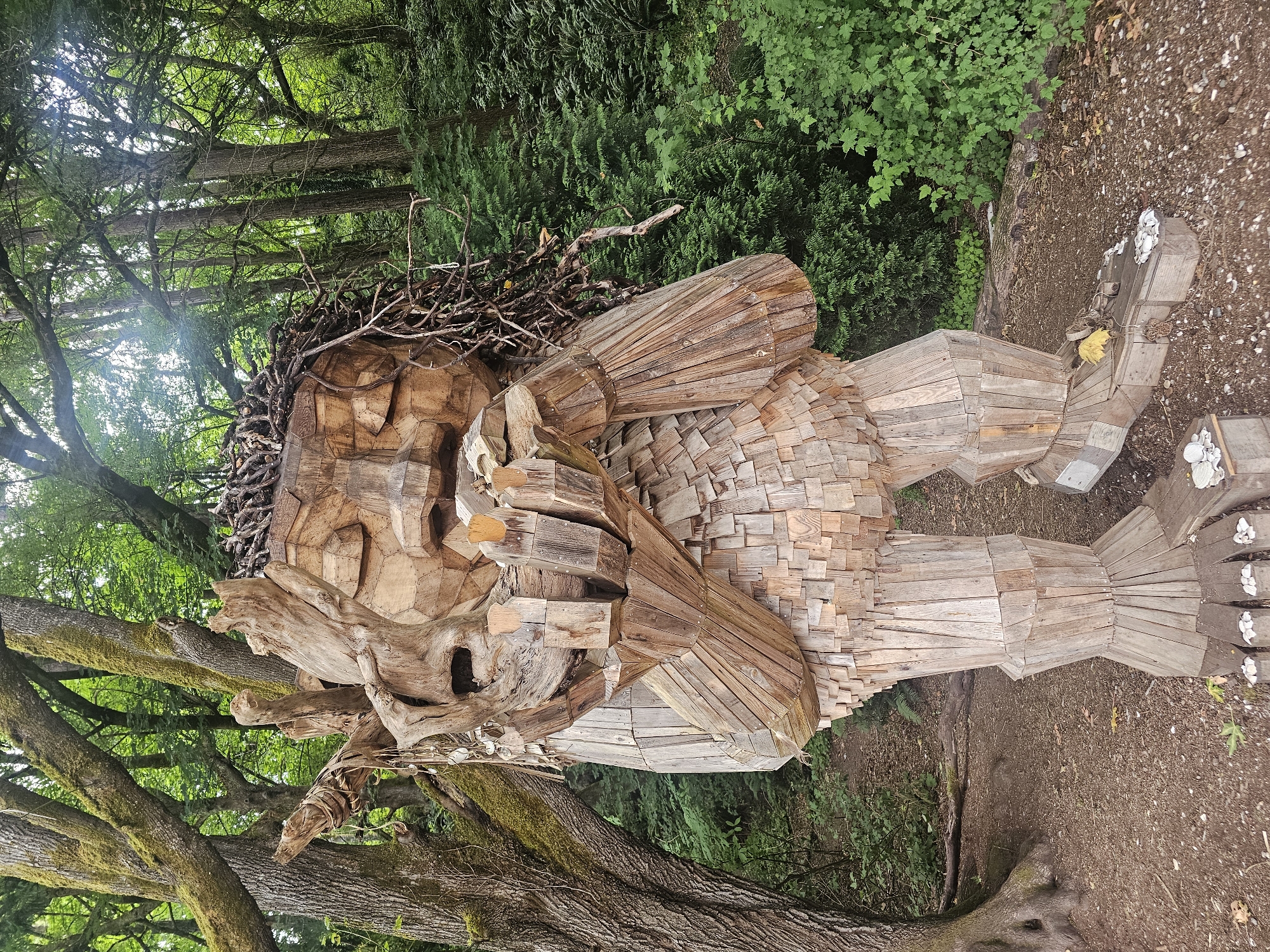 The image depicts a large wooden sculpture of a troll in a forest setting. The troll is made of many wooden planks and pieces, giving it a textured appearance. Its arms are raised and holding a piece of wood or stone to its face, and its eyes are closed. The troll's hair appears to be made of twigs and branches. The sculpture is situated on a dirt path surrounded by lush green trees and foliage. In the foreground, at the troll's feet, are several small white stones. The overall scene is one of an outdoor art installation within a natural environment. This sculpture is located in the Ballard neighborhood of Seattle, Washington, United States, specifically in the Golden Gardens Park.