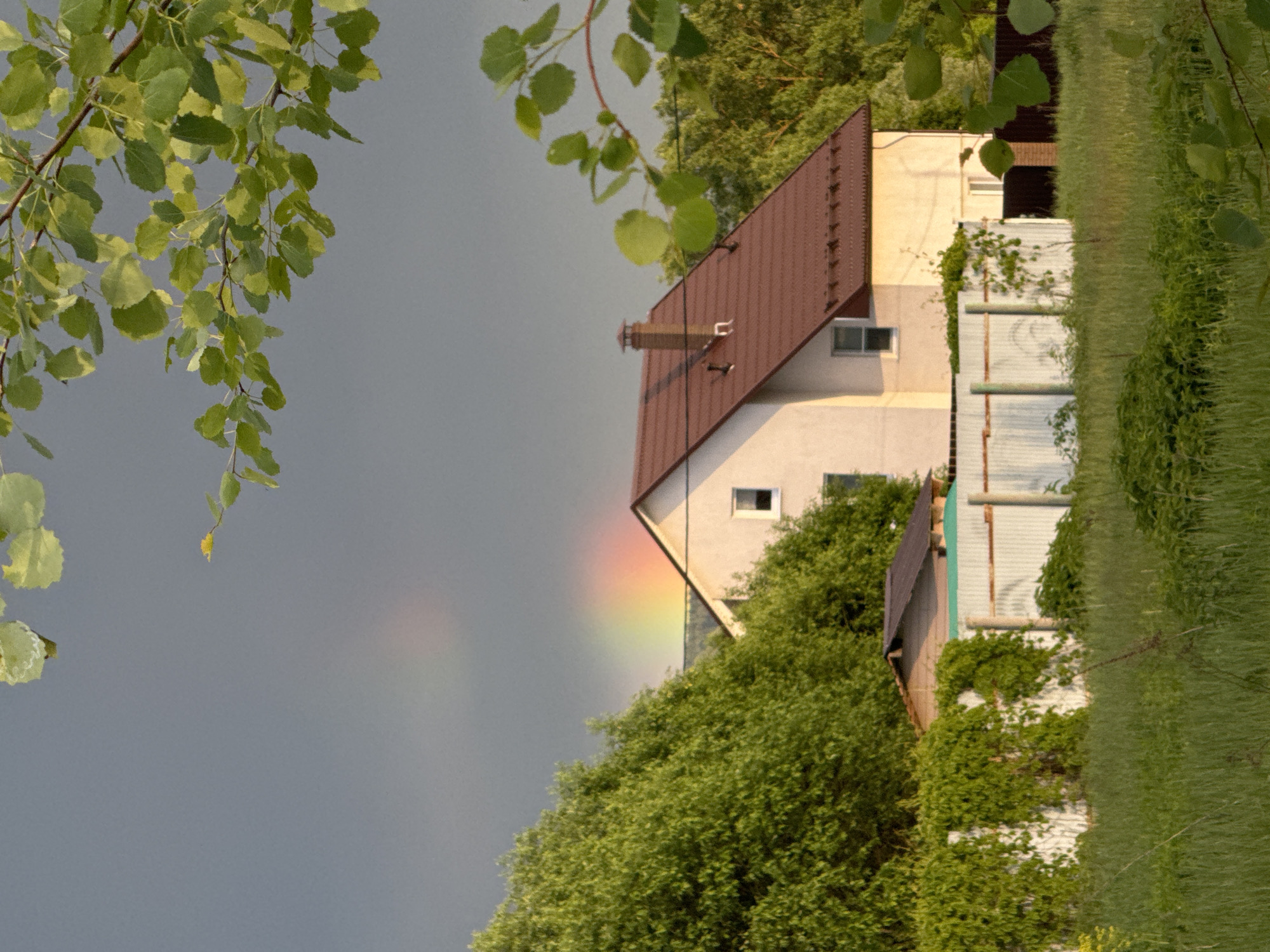 The image depicts a two-story house with a brown roof, situated amidst lush greenery. A faint rainbow is visible in the sky behind the house. Branches with green leaves frame the image on the left. The house is partially obscured by trees and bushes. A fence runs along the right side of the house. The image may be related to the city of Vysokinichi, Russia, though specific landmarks or contextual elements confirming the location are absent.