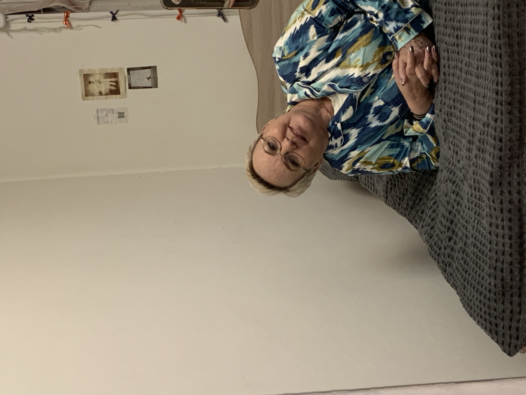 An older woman with short, light-colored hair and glasses is seated indoors, likely on a bed or sofa, in Chaikovsky, Russia. She wears a patterned long-sleeved shirt with shades of blue, green, and white, featuring an abstract or ikat-like design. Her hands are clasped together, resting on a dark grey, textured blanket that covers the seating surface. Her fingernails are manicured, possibly with a metallic polish. She has a gentle smile and is looking towards the viewer, appearing relaxed.
The background is a plain, light-colored wall. On the wall, above and behind her, are several small items. To the left, a sepia-toned photograph of a standing person is visible. To its right, a smaller, darker picture, possibly another photo or drawing, is displayed. Below these, a small white piece of paper with faint, illegible text is attached. Near the top edge of the wall, small decorative bows in red and blue are affixed to a string or wire. The lighting suggests an indoor setting during the daytime. No clearly legible text is visible in the image.