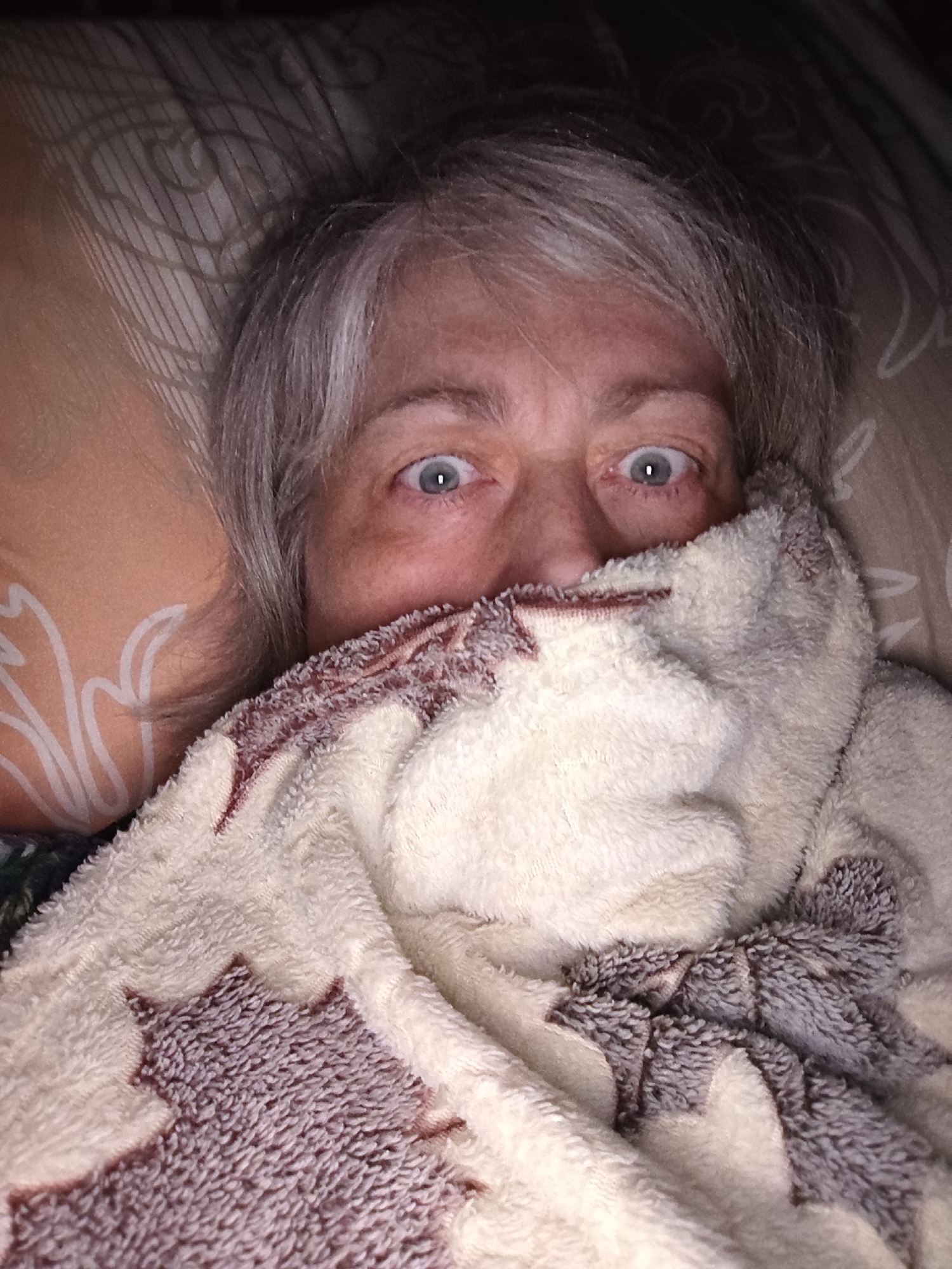 A close-up, overhead shot shows a person with gray hair and light-colored eyes looking directly at the camera, partially covered by a white and brown patterned blanket. The person is lying down, with part of their face obscured by the blanket. The background consists of pillows with a subtle floral pattern. There is no identifiable information about the location in Krasnodar, Russia.