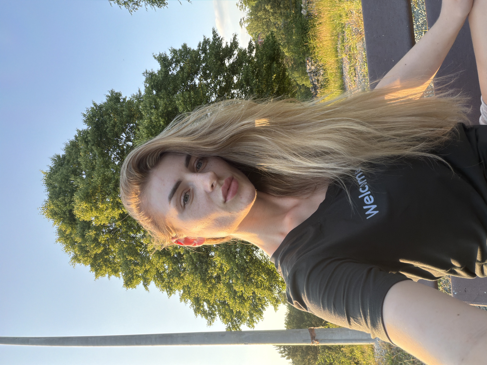 A medium-shot, outdoor selfie features a young Caucasian woman with long blonde hair and fair skin, sitting on a bench in what appears to be a park or natural setting. The golden hour sun illuminates her face from the right, casting soft highlights and shadows. She is wearing a dark t-shirt with the word "Welcome" in white text. In the background, a large, leafy green tree dominates the scene, with a clear blue sky above. To the left, a tall, gray pole is visible. The ground around the bench is covered in dry grass and a stone wall.