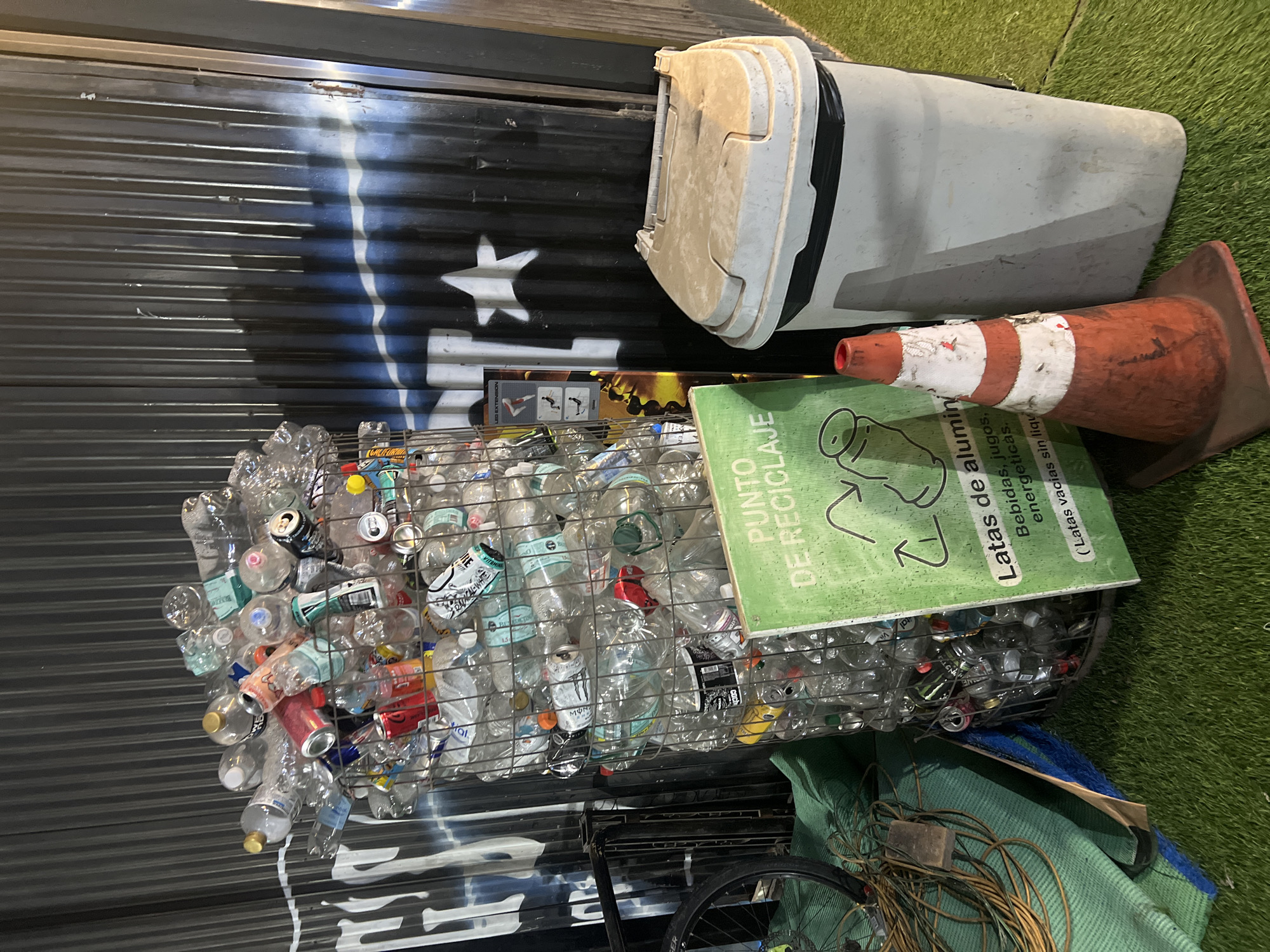 The image depicts a recycling point, likely in Arica, Chile, considering the Spanish signage. A wire cage is overflowing with empty plastic bottles and aluminum cans. On top of the cage rests a green sign with white text, indicating it's a recycling point for aluminum cans ("Latas de aluminio"). Text on the sign specifies the recyclable items include beverages, juices, and energy drinks. A grey trash can with a black rim sits on top of the painted corrugated metal wall behind the recycling cage. An orange traffic cone with a white stripe lies next to the trash can. A bicycle wheel is partially visible in the bottom left corner, amidst some folded green material and a coiled electrical cord. The painted corrugated metal wall has a stylized blue and white paint design. Green artificial turf lies to the right.