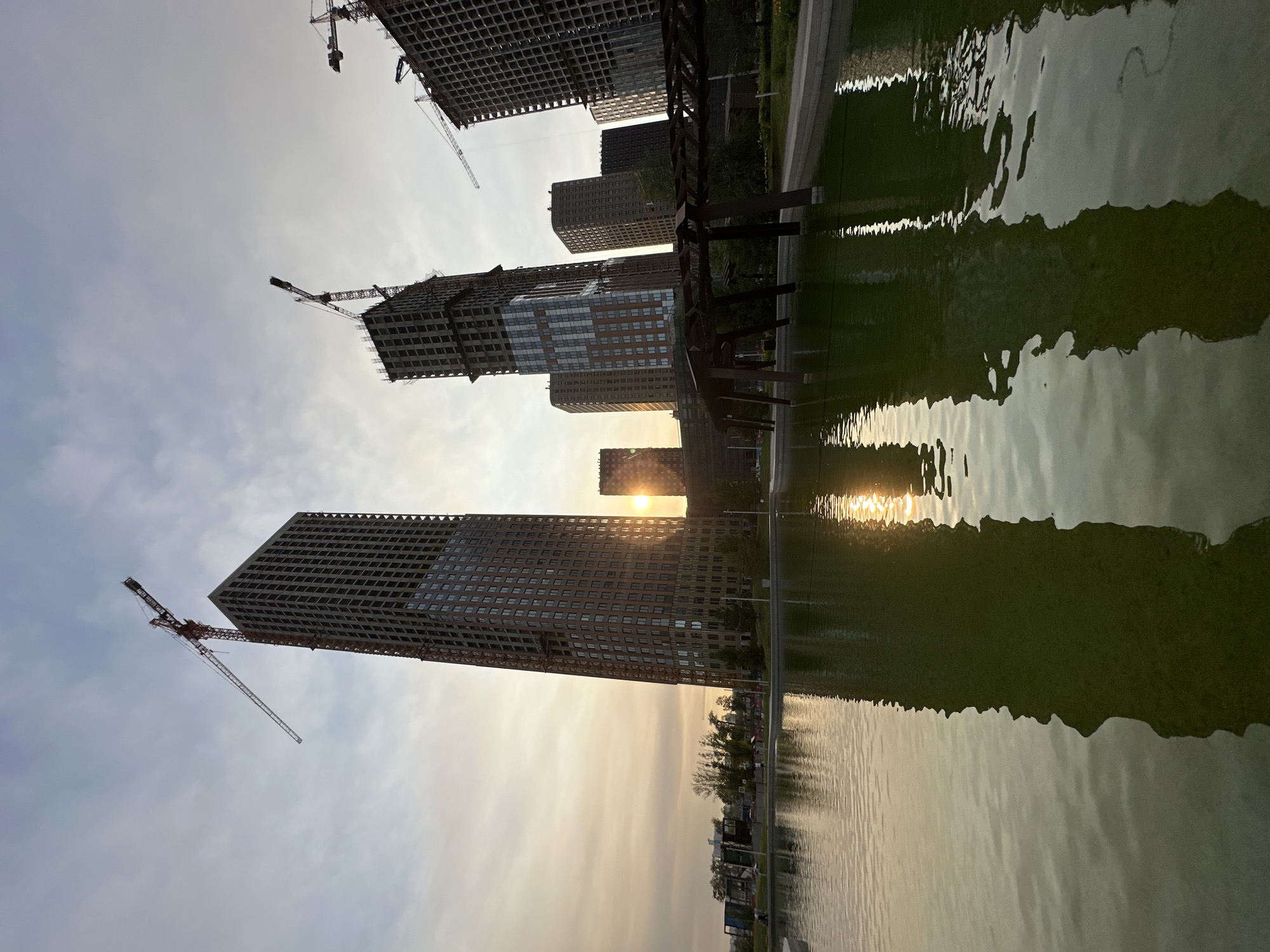 This is a low-angle, wide-shot photograph of a park with a body of water in the foreground, reflecting skyscrapers and construction cranes under a hazy, setting sun. The water, with a greenish tint, occupies the lower half of the frame. A dark, open-lattice structure, possibly a pergola or bridge, spans across the water in the mid-ground, its silhouette sharp against the brighter sky.
Behind the water and structure, several tall buildings are visible. The most prominent is a dark brown skyscraper on the left, its facade covered in small windows. It is under construction, with a large red and white crane extending from its upper section. To its right and further back, other buildings of varying heights and architectural styles are visible, some also appearing to be under construction with exposed concrete frameworks and cranes. The sun, a bright disc, is partially obscured by one of the buildings, casting a warm, golden light across the scene and creating reflections in the water. The sky is a mix of light blue and grey clouds. In the far background, a few trees and what appears to be a city skyline are faintly visible. A few indistinct figures can be seen walking in the park area beyond the water.
This image depicts the "Zaryadye" park in Moscow, Russia, which is known for its modern landscaping and its proximity to new residential and commercial developments, some of which are still under construction.