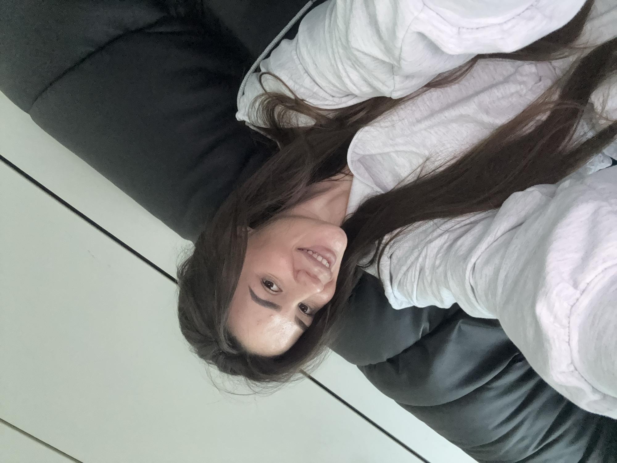 A woman with long, dark brown hair, wearing a light gray hooded sweatshirt, smiles at the camera while lying on a dark gray cushioned surface. She is indoors, with a light-colored wall and a dark vertical line visible in the background. The image is a selfie, taken from a slightly elevated angle.