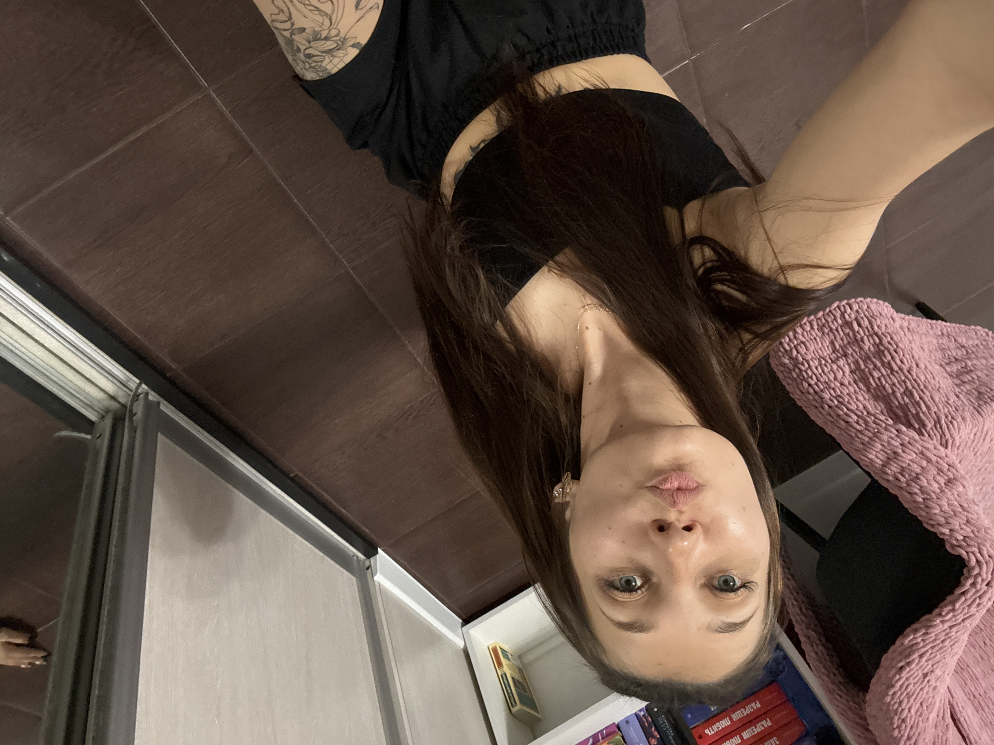 A young woman with long brown hair and blue eyes is sitting on a dark brown tiled floor, facing the camera. She is wearing a black crop top and black shorts, and has a tattoo on her left arm. She is puckering her lips. To her left, a pink knitted blanket is draped over a chair. Behind her, a white bookshelf is partially visible, filled with books. To her right, a sliding mirror wardrobe is present. The room appears to be a bedroom in an apartment in Novokuznetsk, Russia.