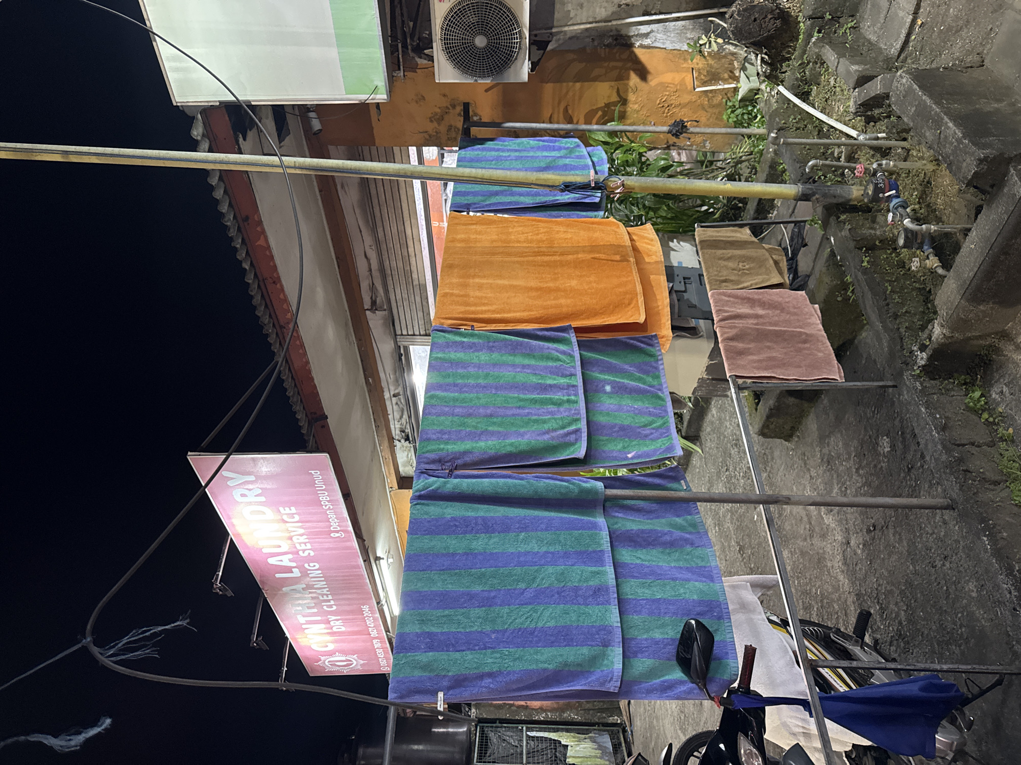 The image depicts a laundry service called "Cyntha Laundry" located in the city of Ungasan, Indonesia. Several towels are hanging on a clothesline to dry outside the laundry shop. The towels are various colors, including blue and green striped, orange, brown, and pink. A motorcycle is parked near the clothesline. The environment appears to be an urban setting with buildings and other structures visible in the background. It is nighttime, and the scene is illuminated by artificial lights.