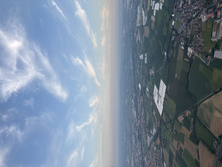 The image displays an aerial panoramic view of a rural and semi-urban landscape, identified as the area surrounding Grumello del Monte, Italy.
The upper third of the frame shows a light blue sky with scattered, elongated white cirrus clouds. The horizon line, appearing hazy and distant, separates the sky from the terrestrial features.
The lower two-thirds of the image depict a vast, largely flat terrain. The ground is characterized by an extensive network of agricultural fields, presenting a mosaic of green and brown hues. Several large, rectangular complexes of white, translucent structures, identified as commercial greenhouses, are prominently visible; significant clusters are located in the mid-left and mid-right portions of the frame. Linear infrastructure, including roads and what appear to be railway lines, crisscrosses the landscape, connecting distinct areas. One major roadway is visible extending diagonally from the lower-left.
Urbanized areas are present as clusters of buildings with predominantly reddish-brown roofs, consistent with Italian towns or villages. A notable urban cluster in the lower-right quadrant includes a sports stadium. Additional industrial or commercial buildings with large, flat roofs are interspersed within or adjacent to these settlements. No human figures, vehicles, or specific actions are discernible within the static scene.