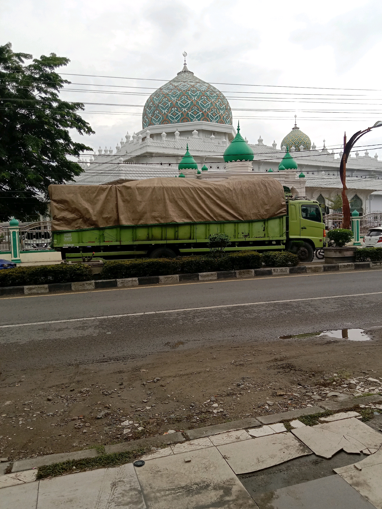 The image depicts the exterior of a mosque in Lhokseumawe, Indonesia, with a large green truck parked in front of it. The mosque features a prominent, ornately decorated dome with teal and orange patterns, and several smaller green domes. White architectural elements and decorative railings are visible. In the foreground, a green tarp-covered truck with a visible "HDC SMS" logo on its side is parked parallel to the road. The truck appears to be a cargo truck with a large covered load. The road in front of the truck shows signs of wear, with patches of dirt, gravel, and a puddle of water. A sidewalk with cracked tiles is visible in the immediate foreground. A large tree with dark green foliage is present on the left side of the image, partially obscuring the mosque. Utility lines are visible in the sky. The sky is overcast with a light gray hue. The overall scene suggests a street view in an urban environment with a prominent religious building and a commercial vehicle.