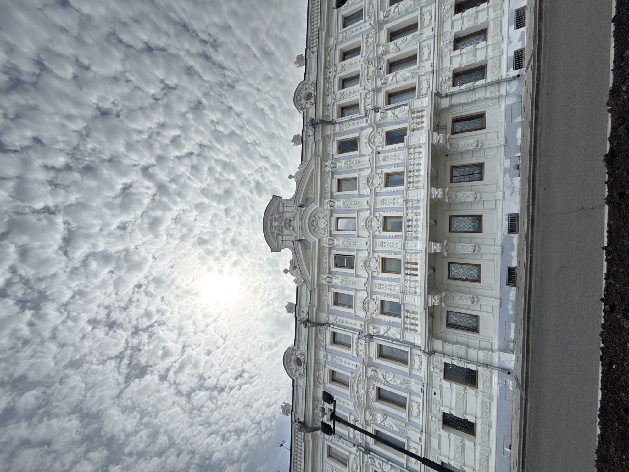The image presents a low-angle view of a building with ornate white and lavender facade details against a sky filled with altocumulus clouds. The sun is visible through the clouds. The building has multiple windows with dark frames. A streetlight pole is in the foreground. The edge of a street or sidewalk is visible on the right side of the frame. The building is believed to be the Rukavishnikov Estate in Nizhny Novgorod, Russia.