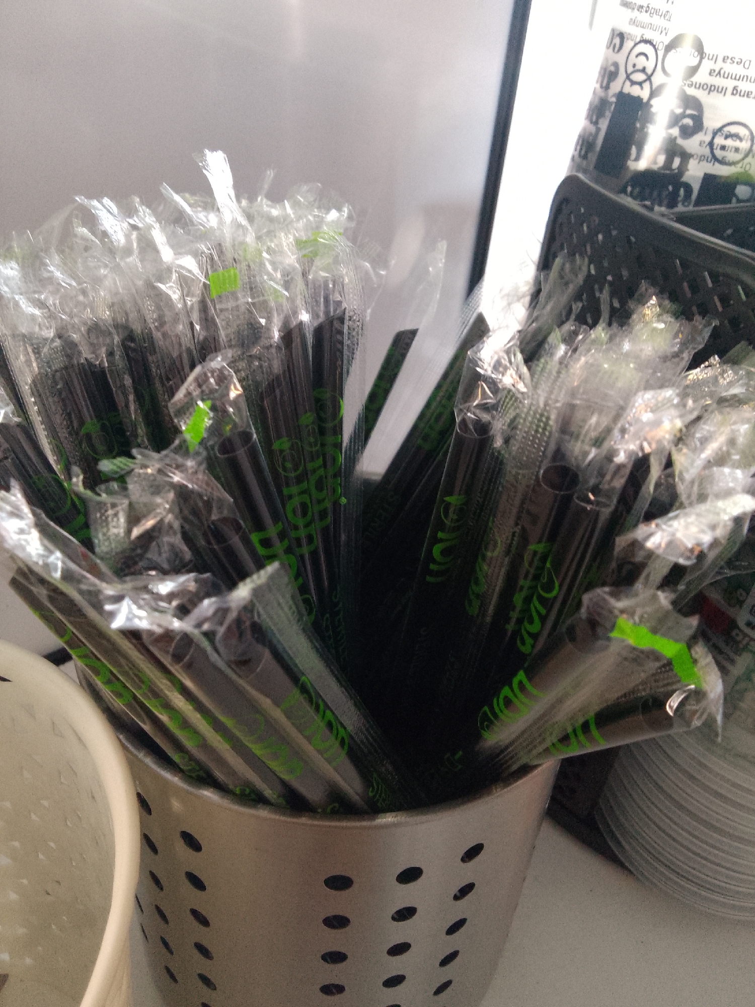 The image shows a close-up of a metal utensil holder filled with individually wrapped black drinking straws. The straws have green lettering on their wrappers, which appears to be a logo or brand name. The holder is metallic and has a pattern of holes. In the background, other items are visible, including stacks of white paper cups and a black plastic container. The overall scene suggests a food service or hospitality setting. The image is taken from a slightly elevated angle, looking down into the holder. There is no indication of the specific location within Trenggalek, Indonesia.