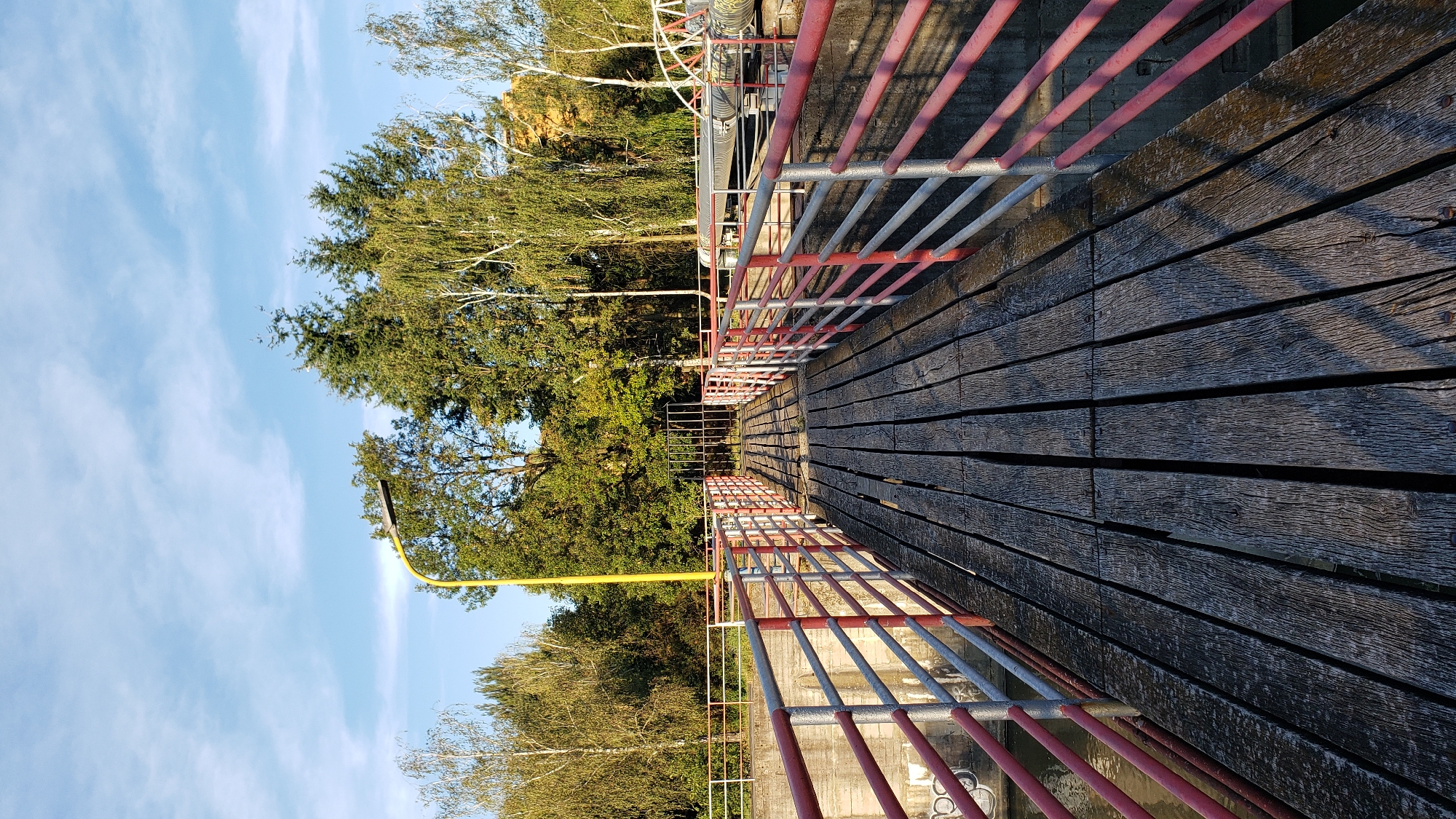 The image depicts a wooden footbridge leading towards a metal gate. The footbridge has weathered wooden planks and is bordered by a railing with red and silver poles and bars. To the left of the bridge, a concrete wall is visible, with some graffiti. The right side of the bridge also features a railing and appears to overlook water. In the background, lush green trees form a backdrop against a clear blue sky with scattered white clouds. A yellow lamppost stands prominently on the left side, partially obscured by trees. The overall scene suggests an outdoor industrial or recreational area, possibly near a body of water. The location is the bridge over the Torysa river in Prešov, Slovakia, part of the city's flood defense system, specifically near the waterworks.