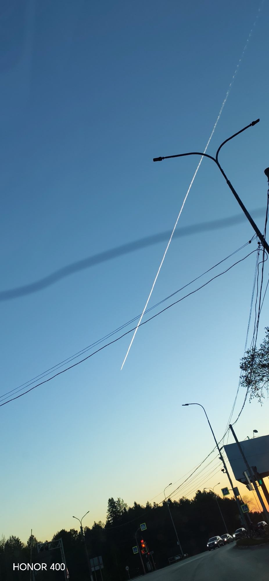 The image shows a contrail in a blue sky, captured in what appears to be the city of Chaikovsky, Russia. The lower part of the frame includes a street with traffic lights, trees, a road, and vehicles. Above, utility poles and wires cut across the view of the sky. The sky gradient transitions from a lighter shade near the horizon to a deeper blue overhead. A long contrail is visible across the sky. The image was taken with a HONOR 400 phone.